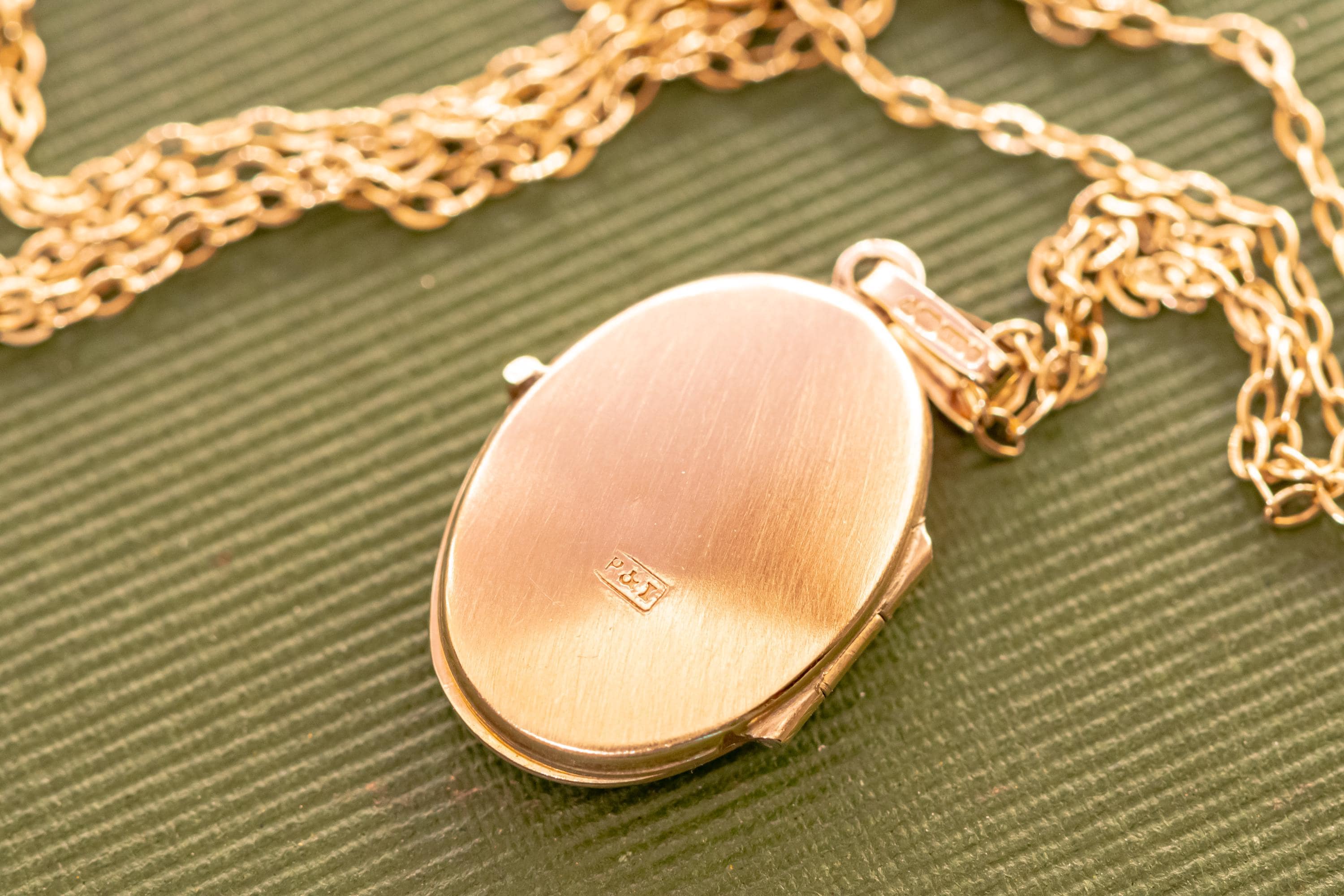 A gold-toned oval locket pendant suspended from a matching link chain. The locket features a brushed metal finish on its front surface with a small rectangular hallmark stamped in the centre and a bail at the top for the chain.