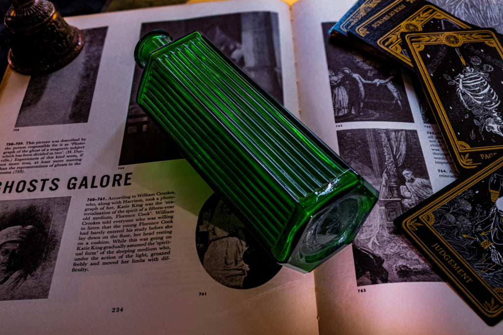 A rectangular, emerald green glass bottle with vertical ribbed detailing along its sides. The bottle features a narrow neck and a flat base, resting diagonally across an open book with printed text and black and white illustrations.