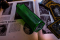 A rectangular, emerald green glass bottle with vertical ribbed detailing along its sides. The bottle features a narrow neck and a flat base, resting diagonally across an open book with printed text and black and white illustrations.
