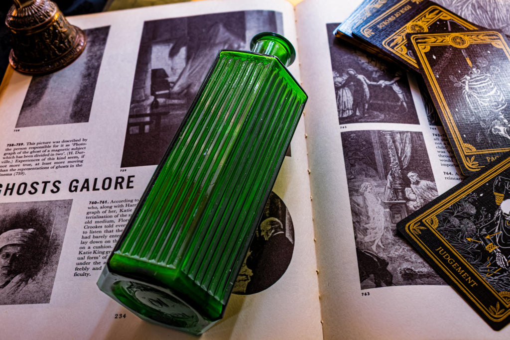 A rectangular emerald green glass bottle with vertical ribbed detailing on its sides. The bottle features a narrow neck and a flat base, resting on an open book with black and white illustrations and the text GHOSTS GALORE.