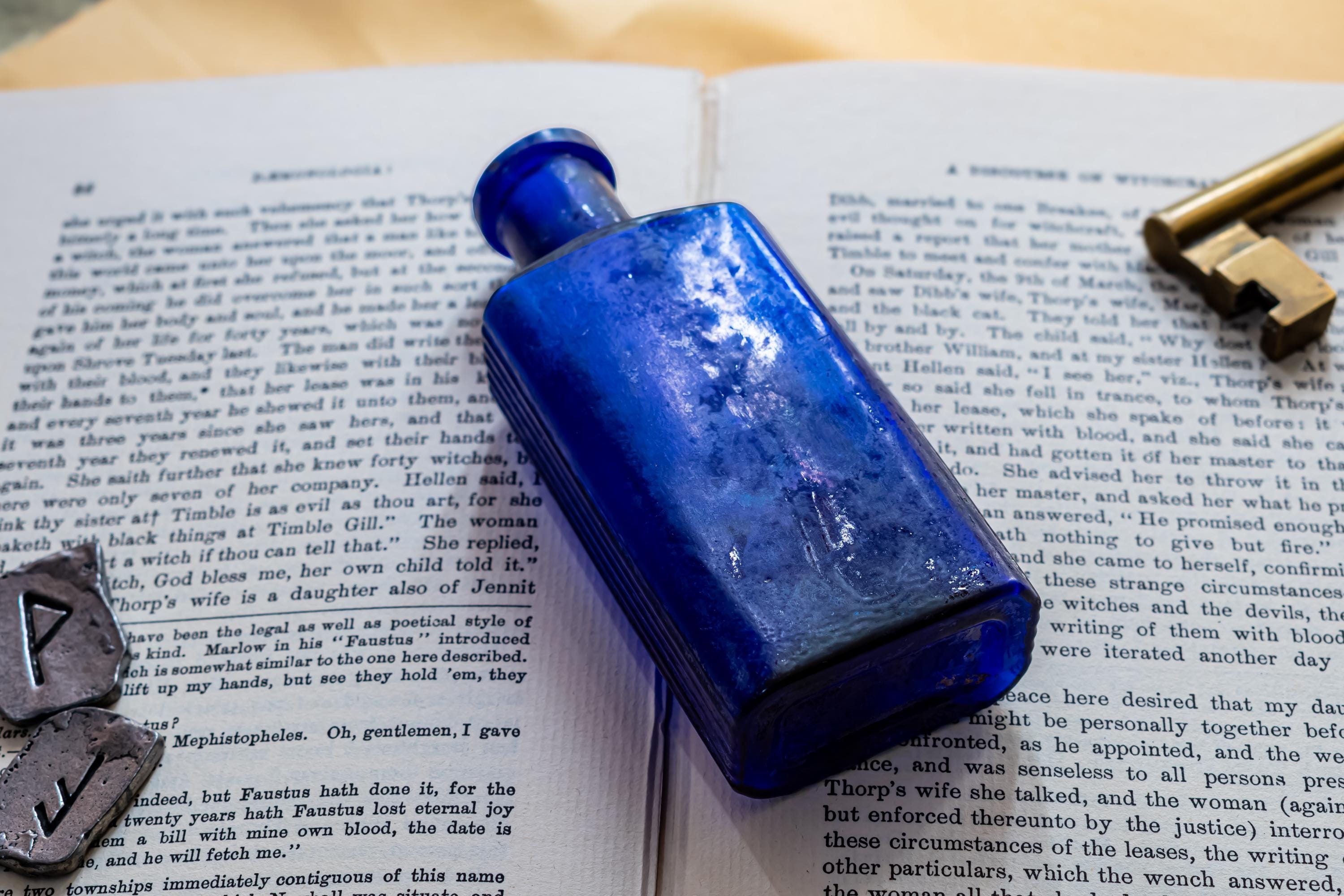 A rectangular cobalt blue glass bottle with a narrow neck, positioned on an open book with printed text. Two small grey stone runes with carved symbols are placed to the left, and a brass-coloured metal key rests on the top right.