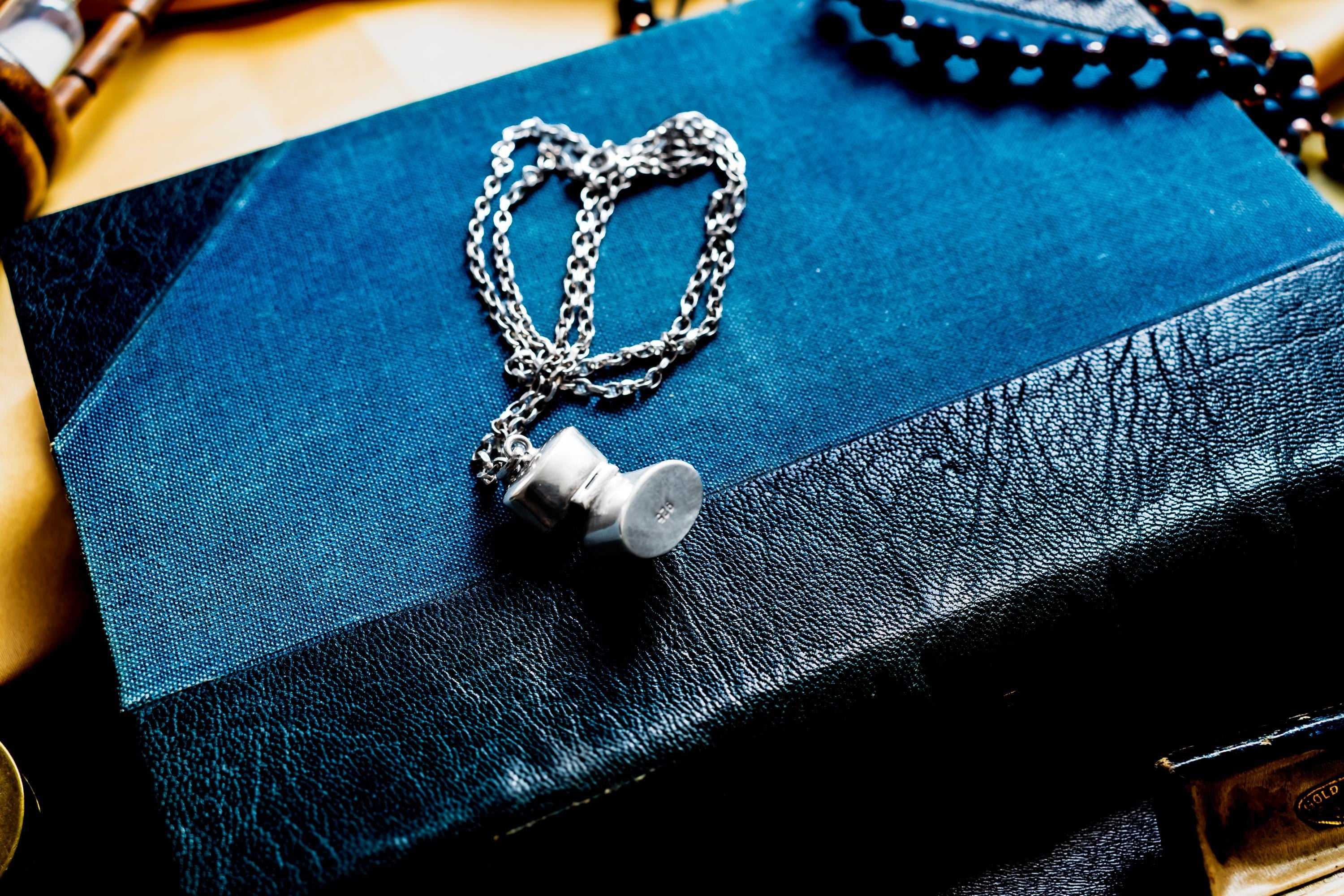 A silver-toned antique silver pill box pendant showing the 925 stamp, suspended from a delicate silver-toned link chain. The pendant rests on a dark blue and black textured book cover, with a portion of a black beaded necklace visible nearby.