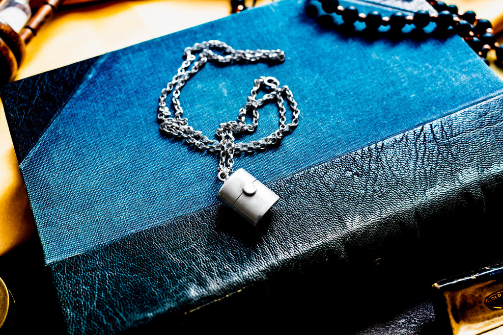 A silver-toned antique silver pill box pendant necklace featuring a small circular clasp detail on the front, suspended from a delicate silver-toned chain. The pendant rests on a textured blue and black book cover with visible grain.