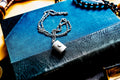 A silver-toned antique silver pill box pendant necklace featuring a small circular clasp detail on the front, suspended from a delicate silver-toned chain. The pendant rests on a textured blue and black book cover with visible grain.