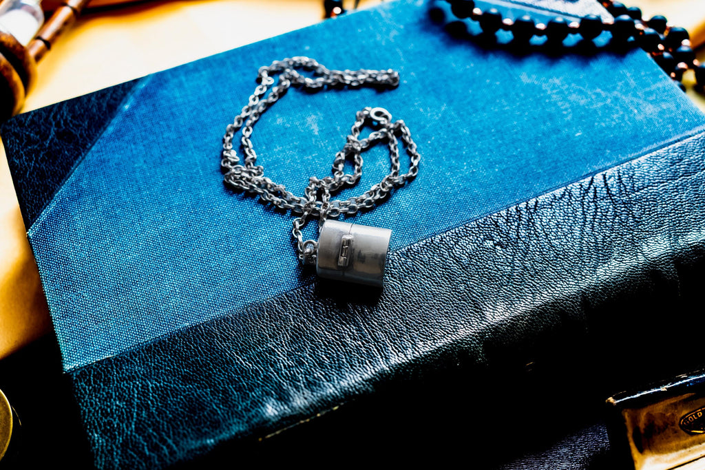 A silver-toned antique silver pill box pendant hangs from a delicate silver chain, resting on a blue book with a textured black leather spine. The pendant features a small rectangular clasp detail on its smooth, metallic surface.