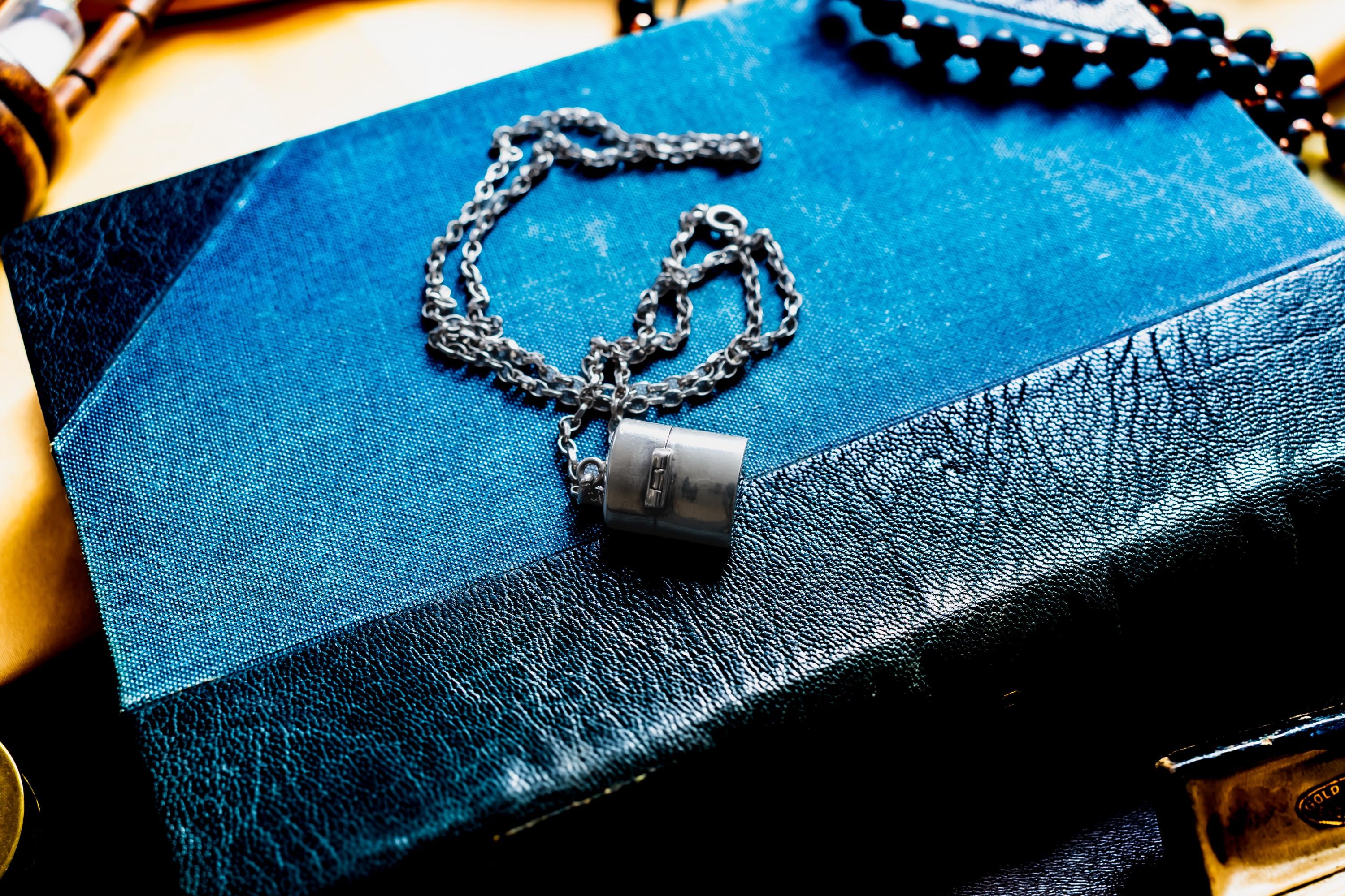 A silver-toned antique silver pill box pendant hangs from a delicate silver chain, resting on a blue book with a textured black leather spine. The pendant features a small rectangular clasp detail on its smooth, metallic surface.