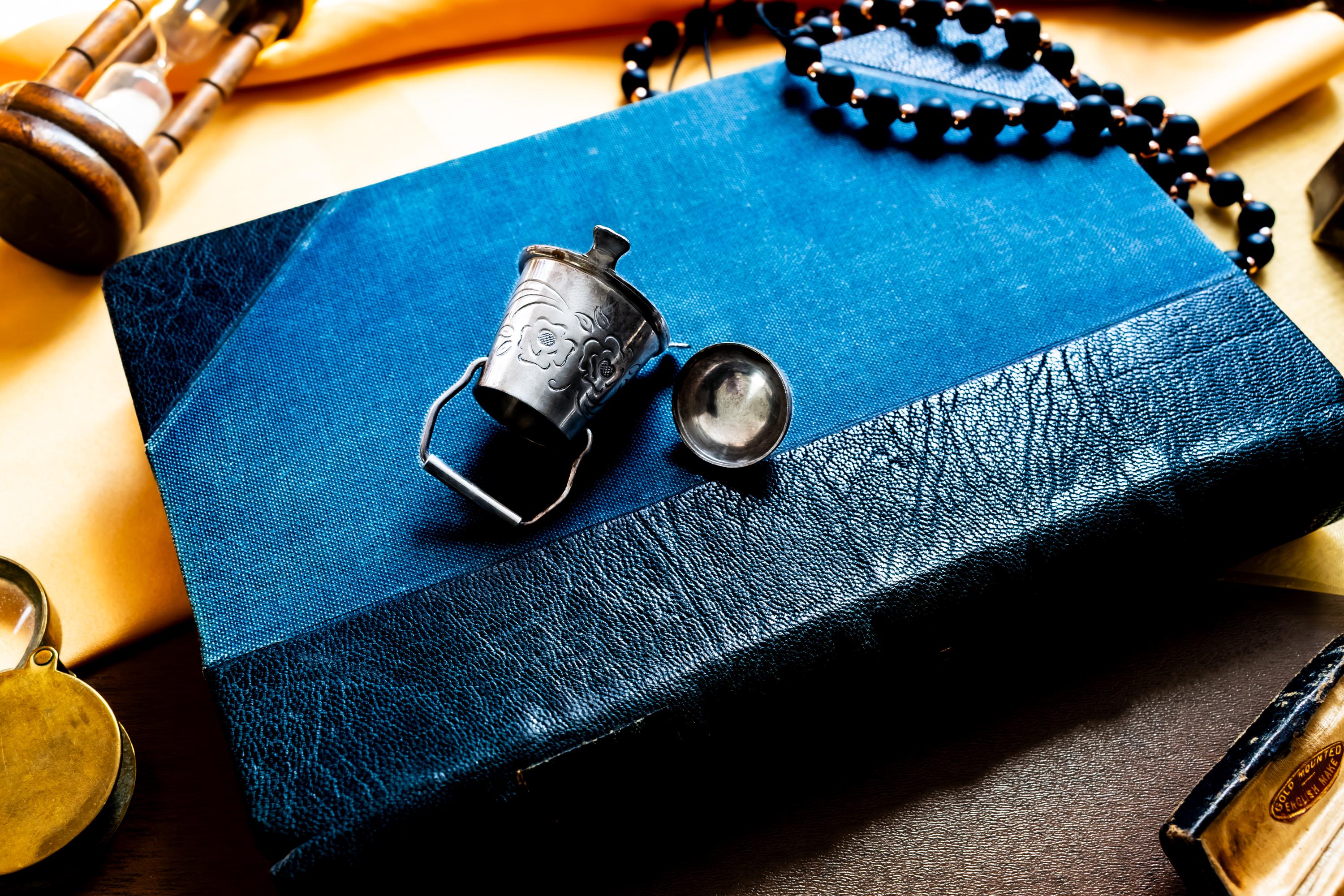 An image showing the side of a Miniature antique silver pail with a small handle, sitting on top of a dark blue vintage book.