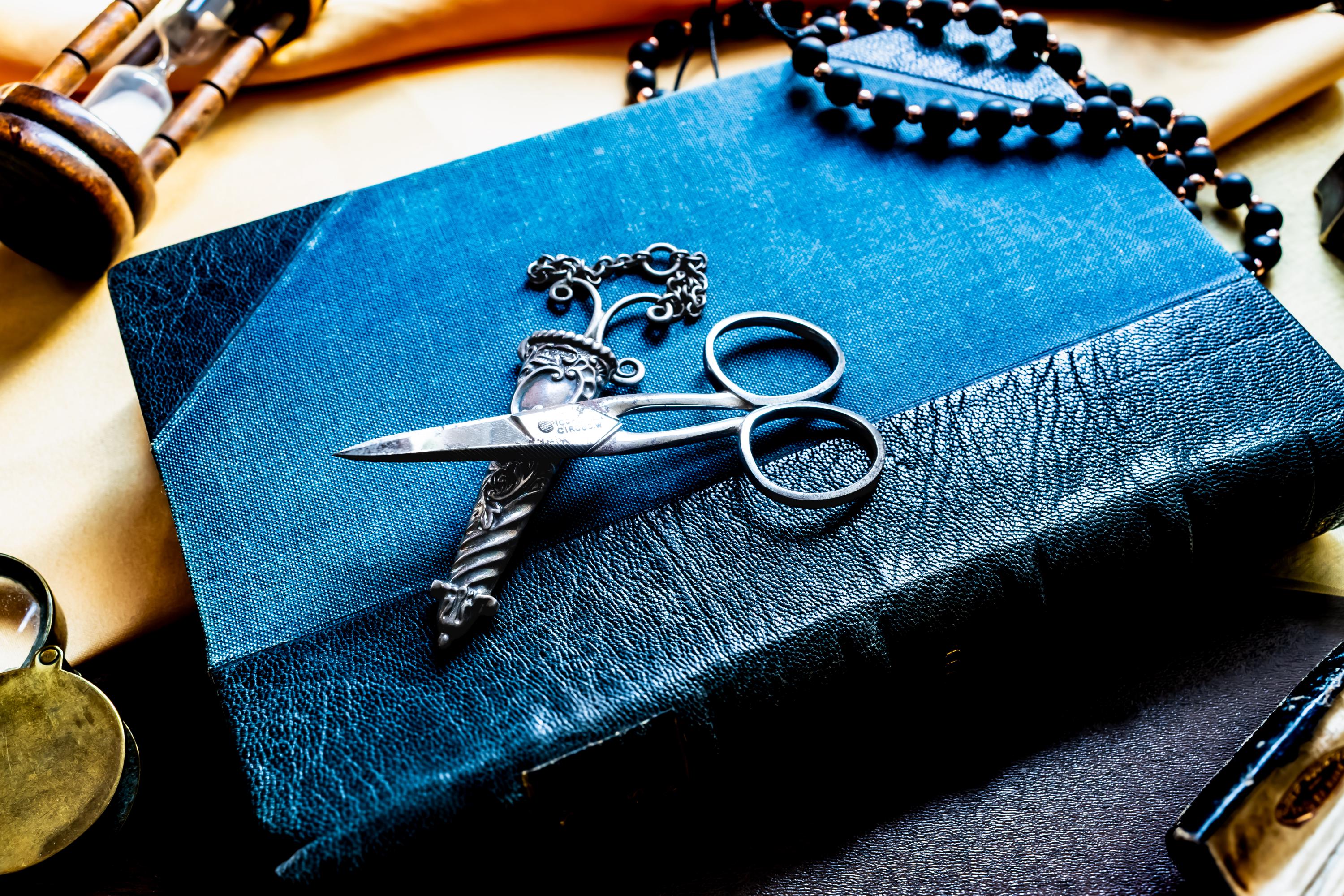 A rectangular book with a textured blue cover featuring a dark blue leather-like spine and edges. Resting on the book is a pair of silver-toned antique metal scissors with ornate, engraved handles and a matching decorative metal sheath with a chain.