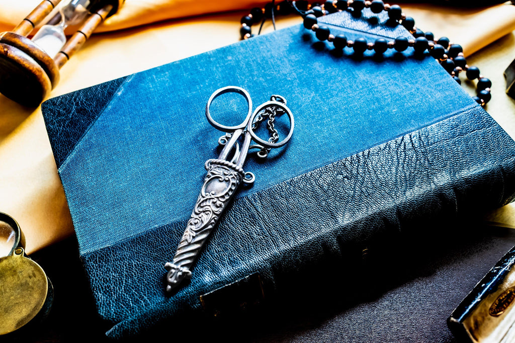 A pair of silver antique scissors with ornate, embossed handles and a matching decorative sheath, resting on a textured blue book. The scissors feature a pointed tip and circular finger loops, with a small chain attached to the handle.