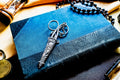 A pair of silver antique scissors with ornate, embossed handles and a matching decorative sheath, resting on a textured blue book. The scissors feature a pointed tip and circular finger loops, with a small chain attached to the handle.