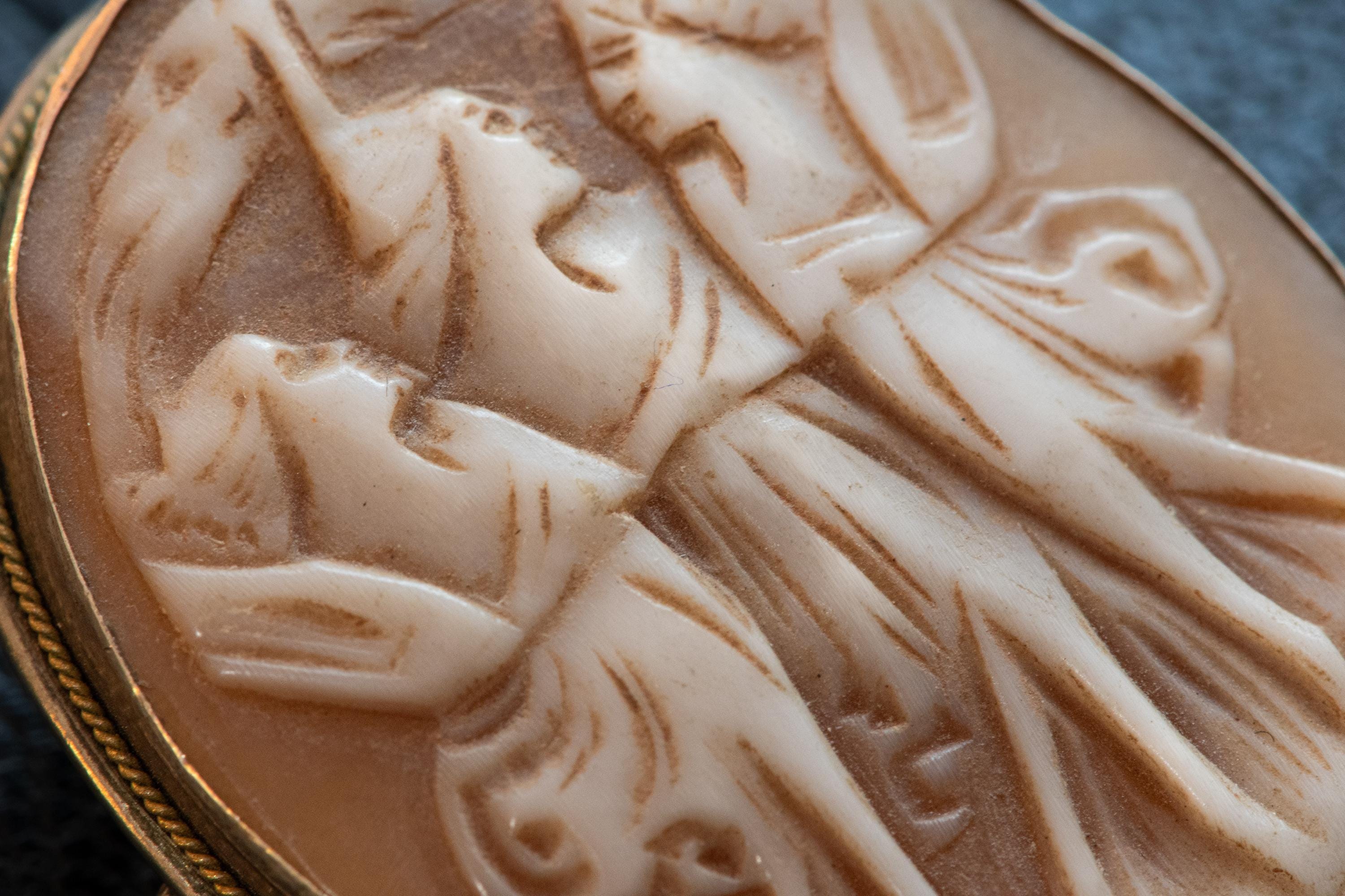 A close shot of an Antique carved shell cameo brooch depicting the Three Graces set in silver, resting on a textured blue background.