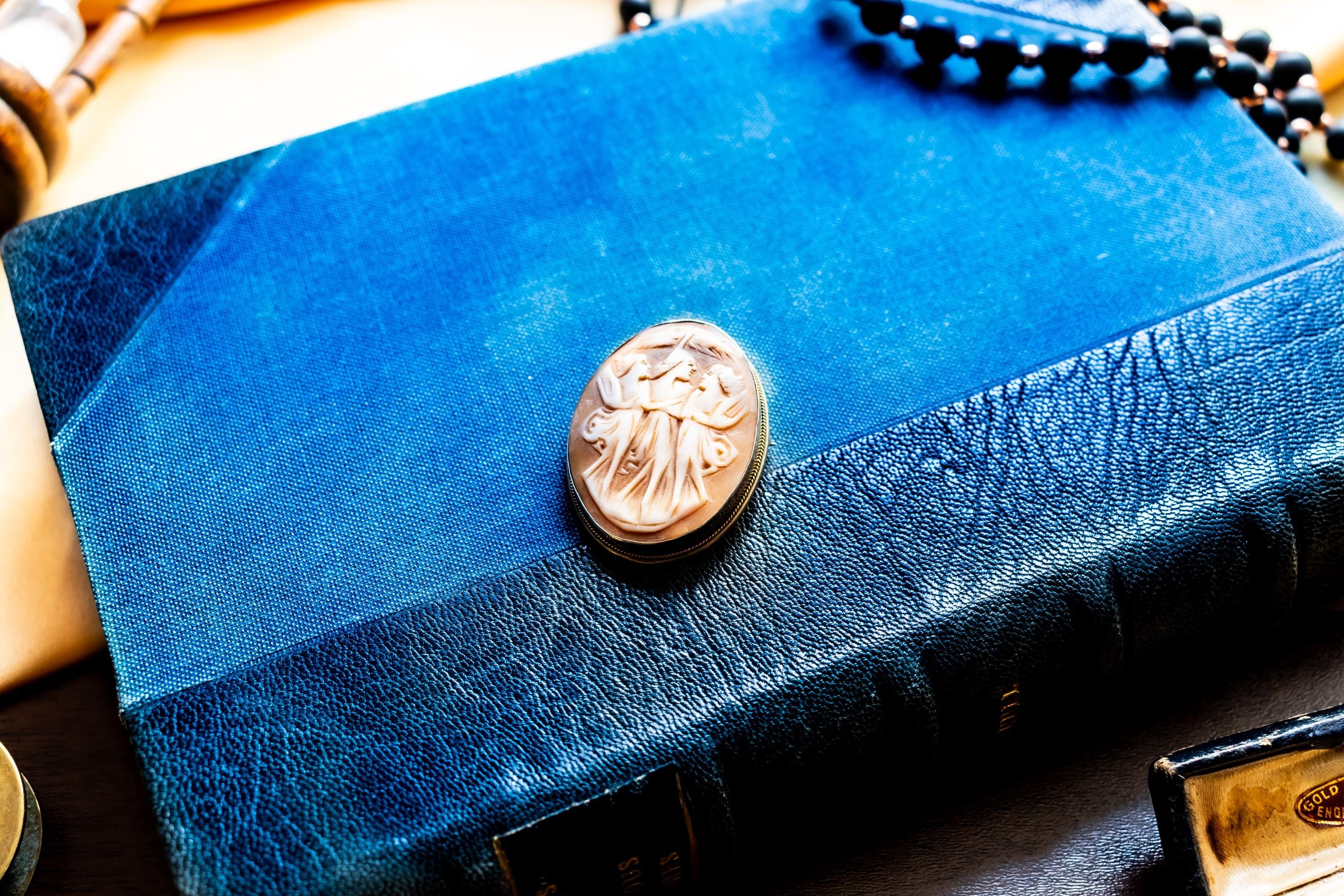 Antique carved shell cameo brooch depicting the Three Graces set in silver, resting on a textured blue background.