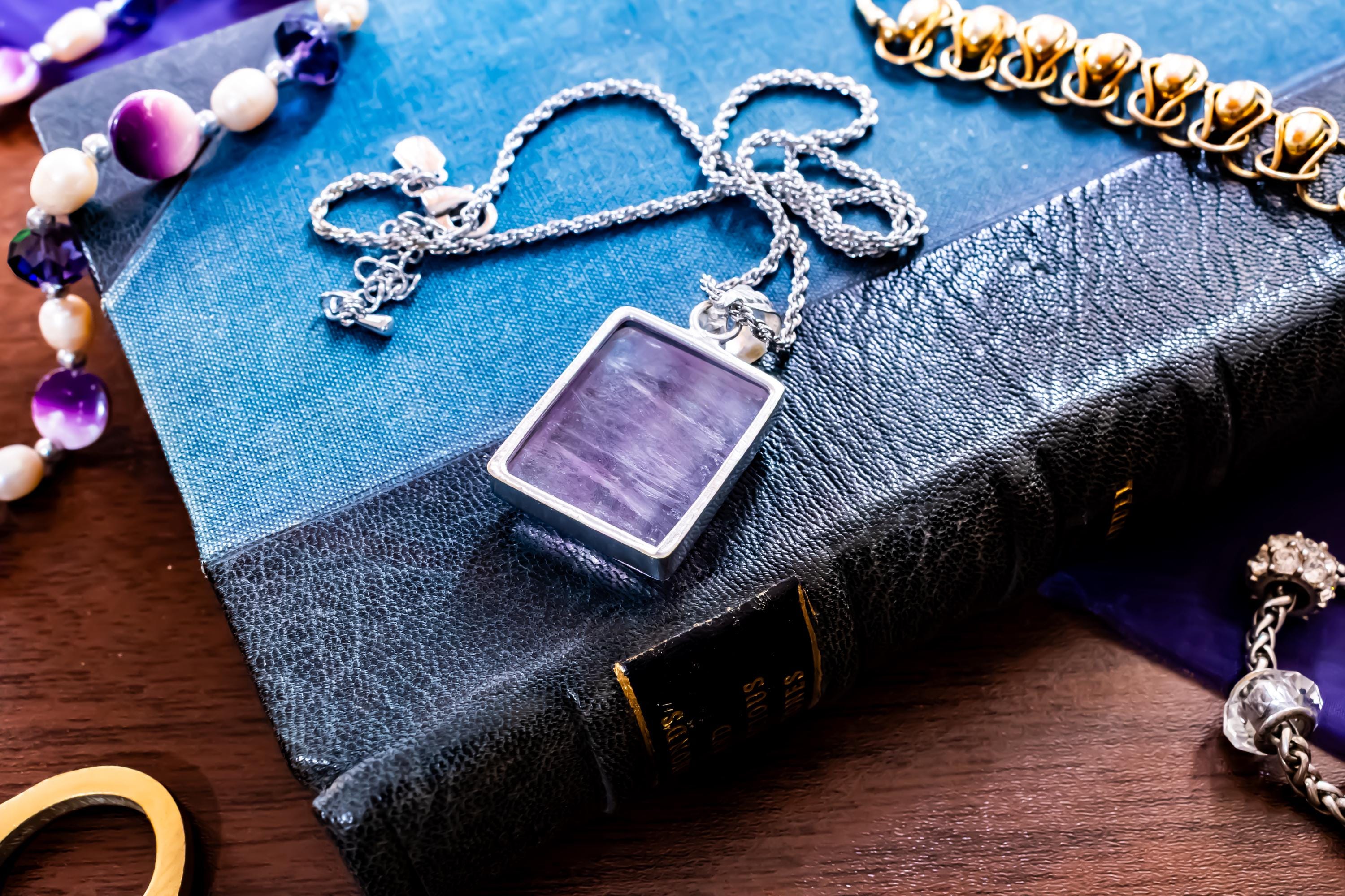 Close-up of a large, rectangular Kunzite gemstone pendant with natural pink and violet banding, set in 925 sterling silver and resting on a textured blue book.
