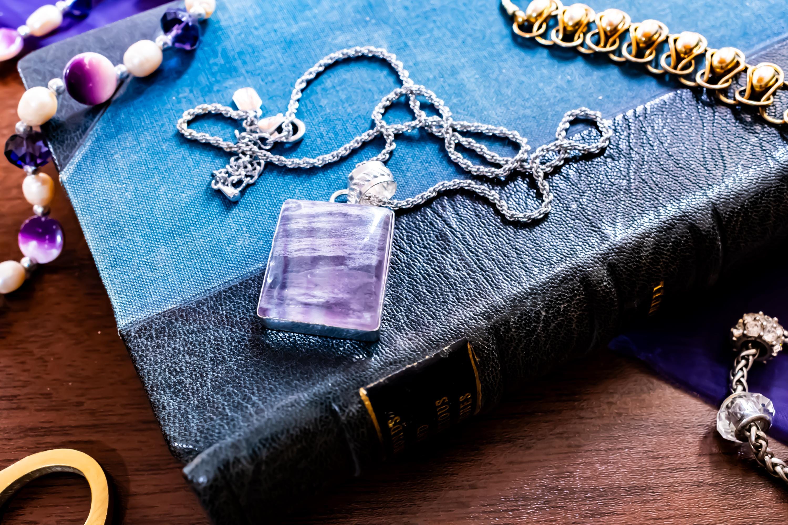 Close-up of a large, rectangular Kunzite gemstone pendant with natural pink and violet banding, set in 925 sterling silver and resting on a textured blue book.