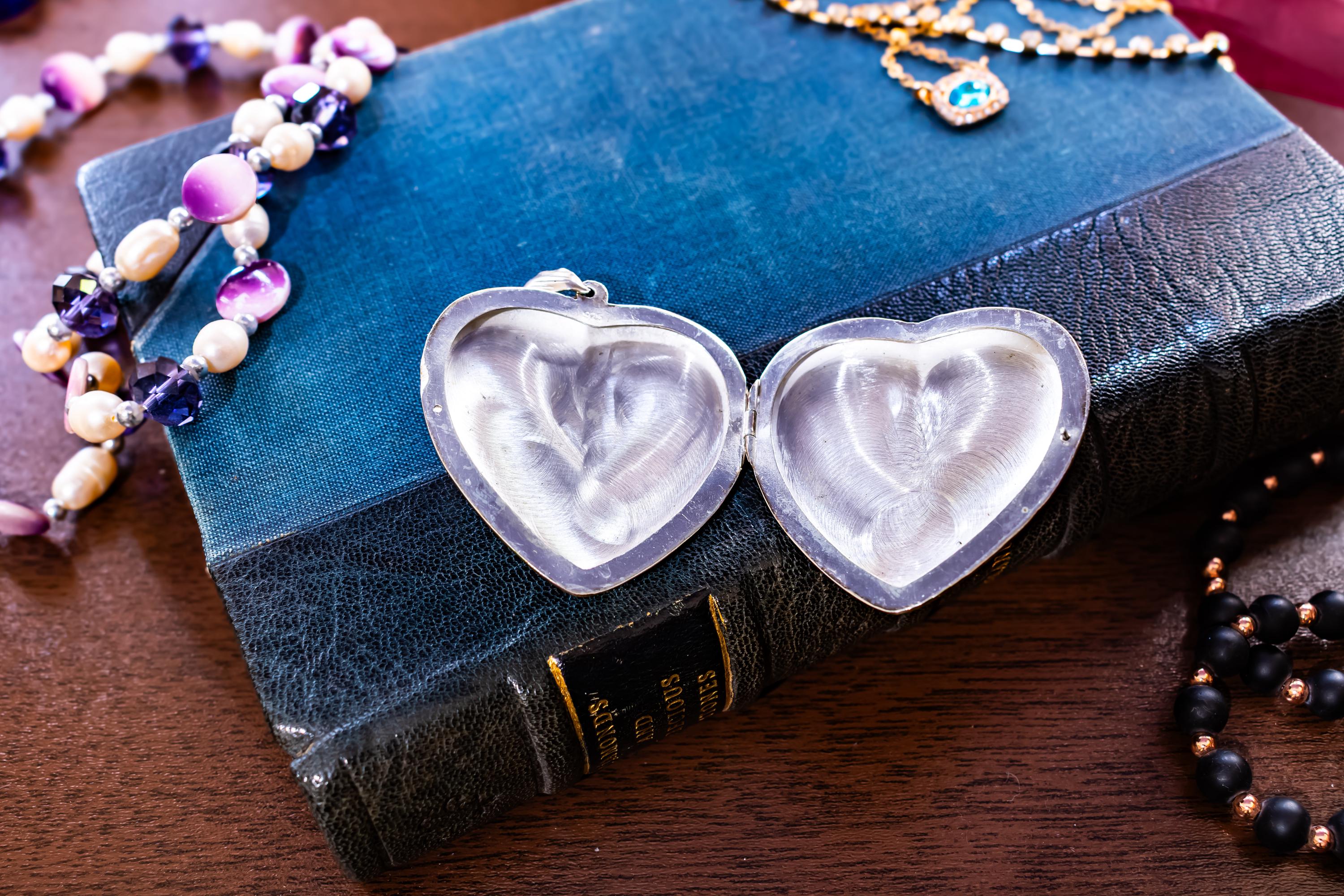 An image of an opened silver-toned heart-shaped vintage locket featuring an intricate engraved floral pattern on its surface. The locket is positioned on top of a textured blue book, surrounded by various beaded necklaces and a gold-toned chain with a blue stone.