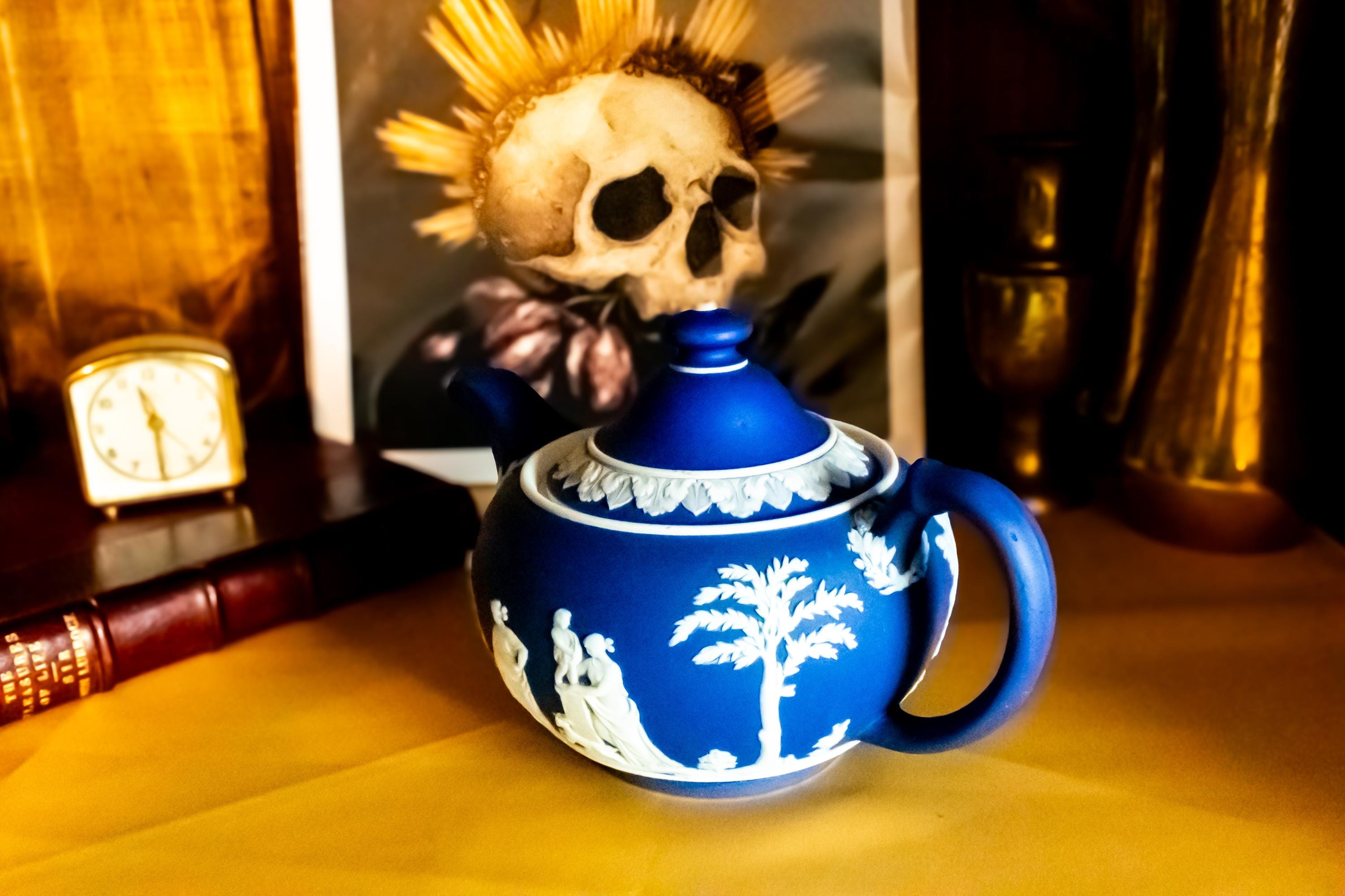 A Matte blue and white team pot resting on a gold material. The teapot has white neoclassical raised images around the pot.