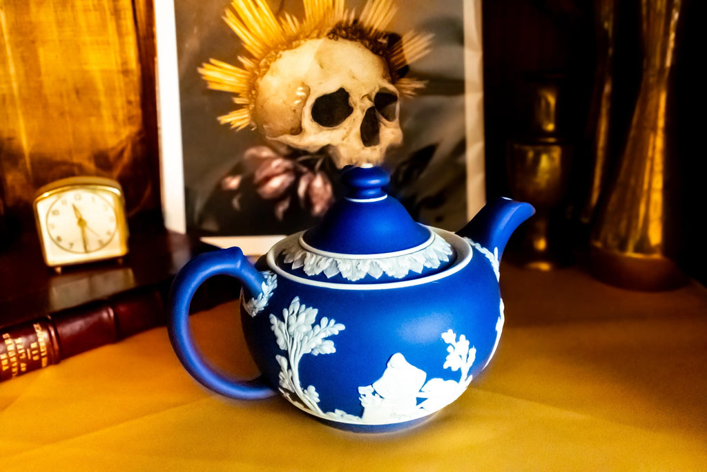 A Matte blue and white team pot resting on a gold material. The teapot has white neoclassical raised images around the pot.