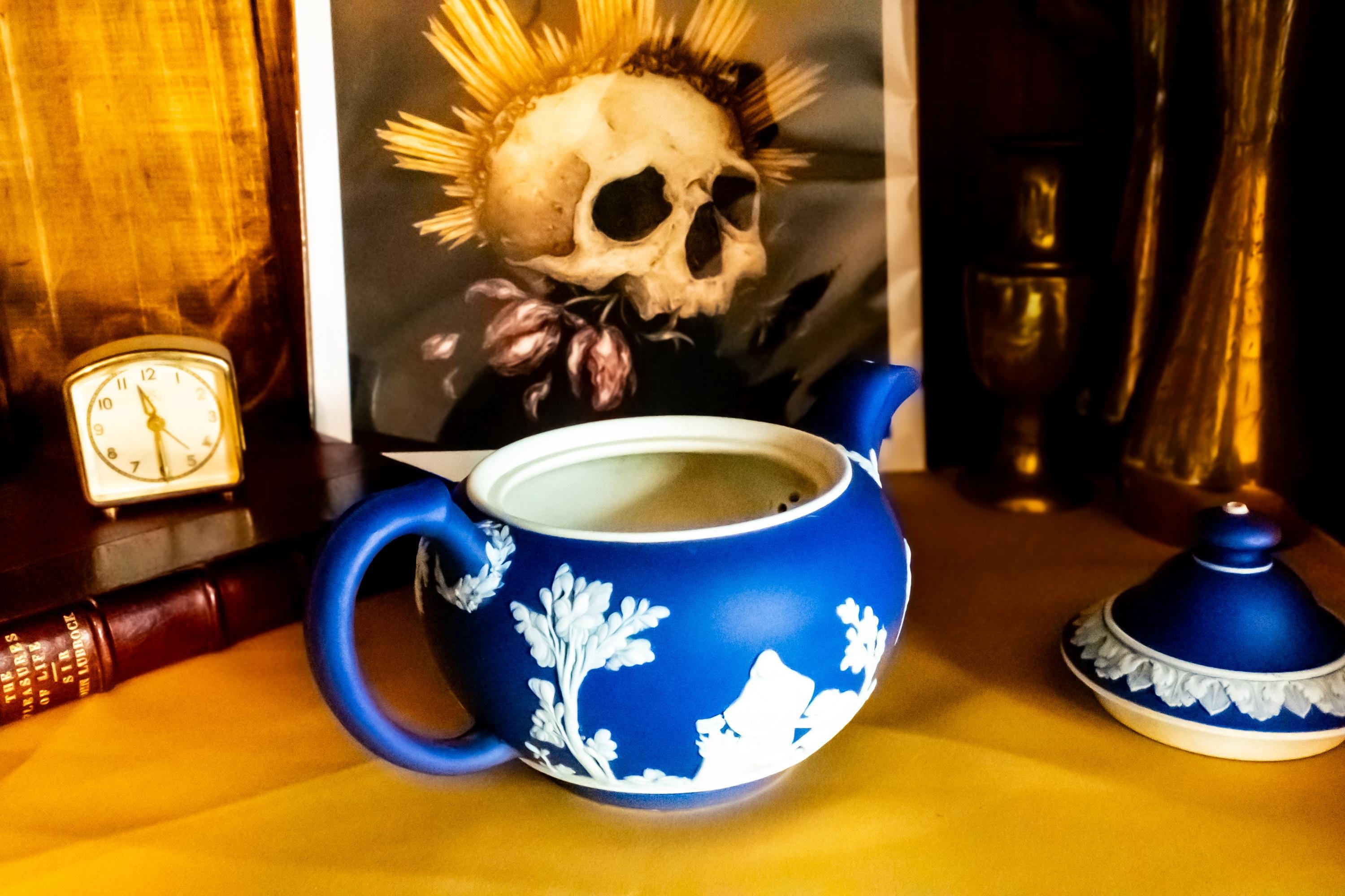A Matte blue and white team pot resting on a gold material. The teapot has white neoclassical raised images around the pot.