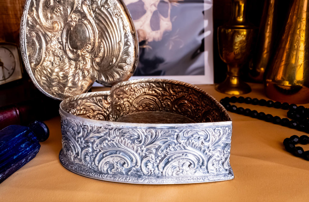 A sterling silver hand engraved jewelry box opened on a shelf with gold material. The jewelry box is in the shape of a heart.