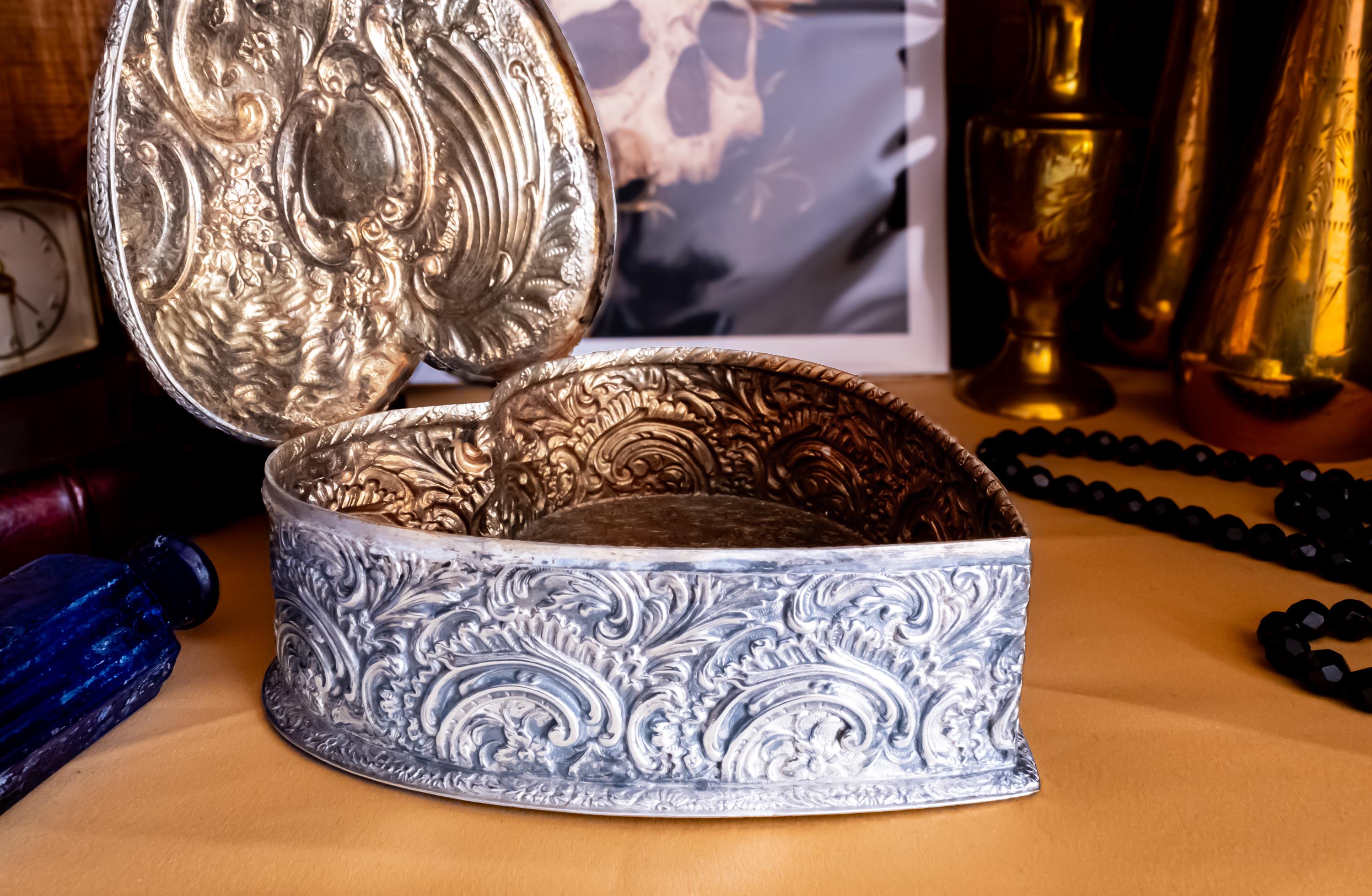 A sterling silver hand engraved jewelry box opened on a shelf with gold material. The jewelry box is in the shape of a heart.