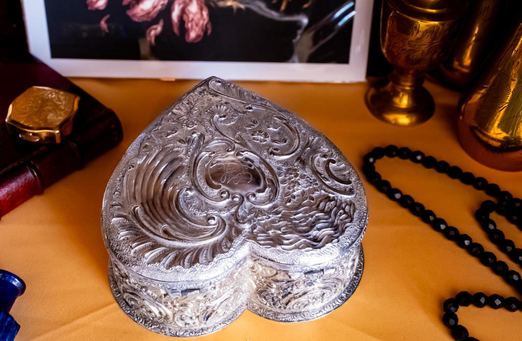 A solid sterling silver hand engraved jewelry box showing the back hinges of the lid. The shape of the jewelry box is a hard. It has engravings on the top and on the sides.