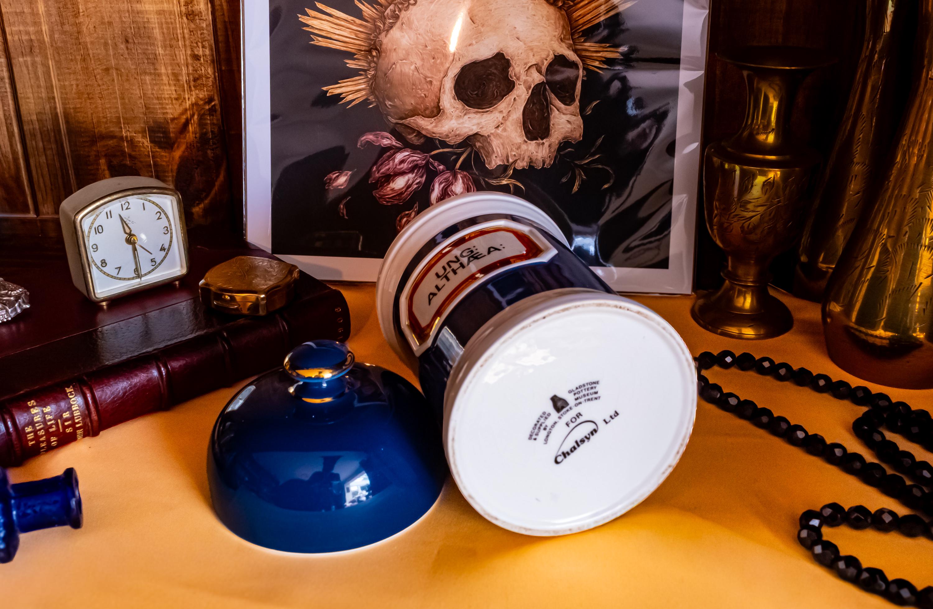 Antique apothecary blue and white jar with a label that says Ung: Althaea. The label is surrounded by gold foil. The white jar has a blue porcelain lid. The jar is laying on its side to showcase the designer label.