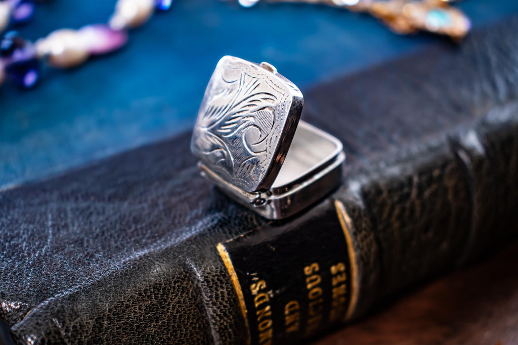 An image of the back of a Tiny square antique silver pill box with engraved floral patterns sitting on a dark textured book
