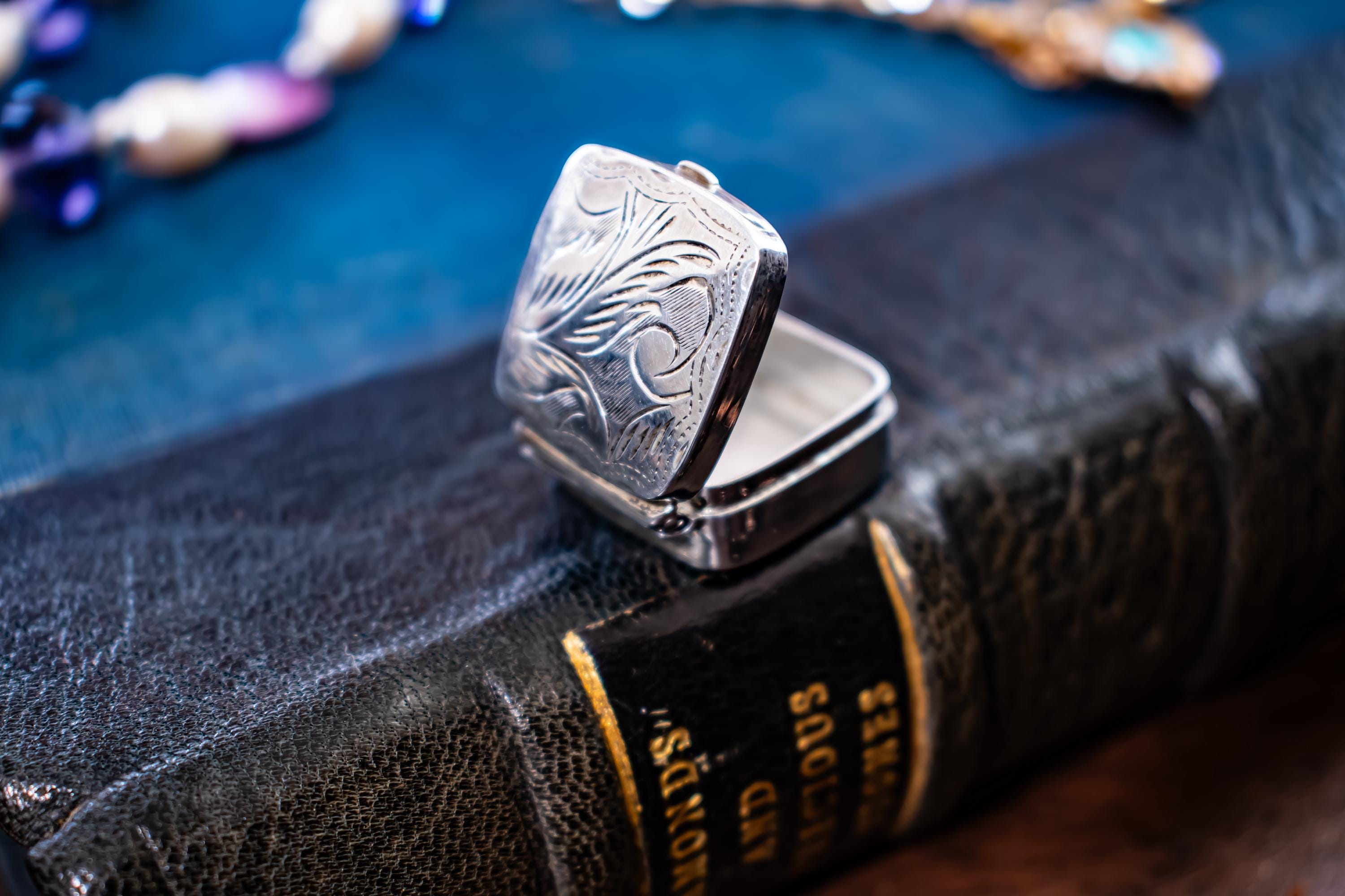 An image of the back of a Tiny square antique silver pill box with engraved floral patterns sitting on a dark textured book