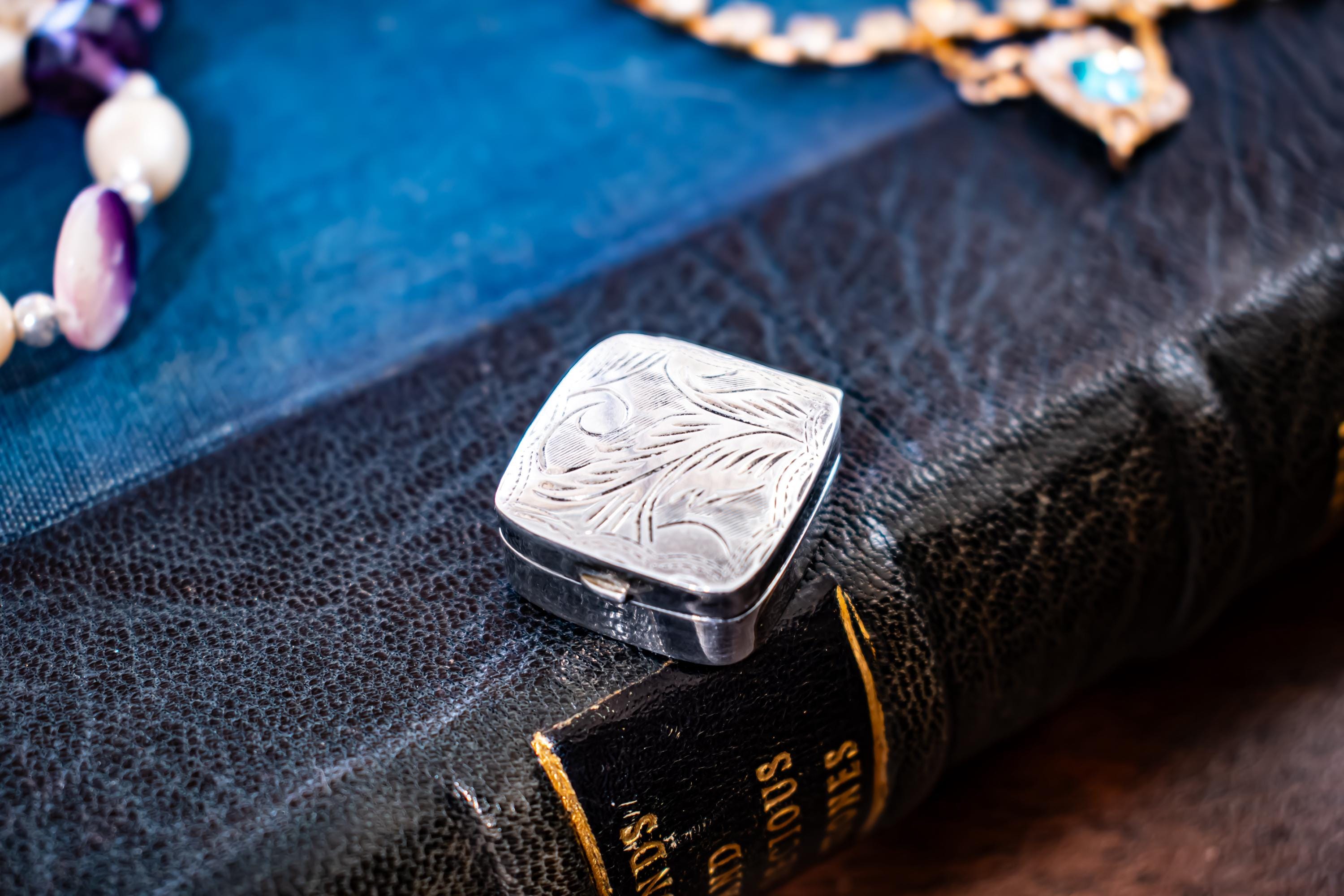 Tiny square antique silver pill box with engraved floral patterns sitting on a dark textured book