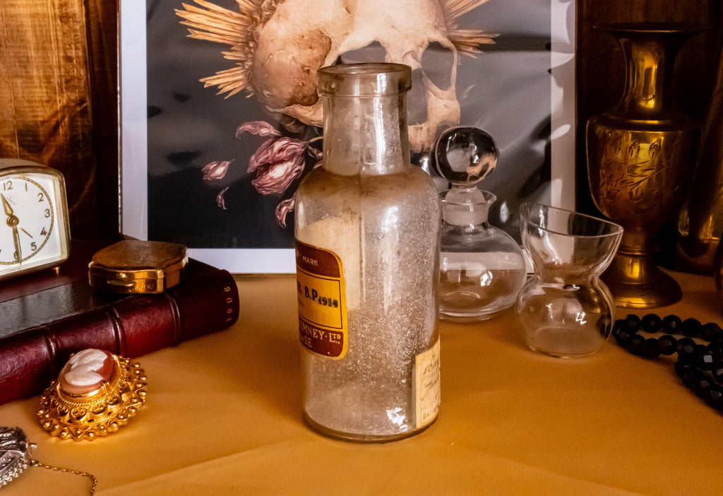 Antique clear glass apothecary bottle with a yellow and red paper label reading "Guaiacol Carb" from London, sitting next to a vintage glass stopper and an anatomical skull print.