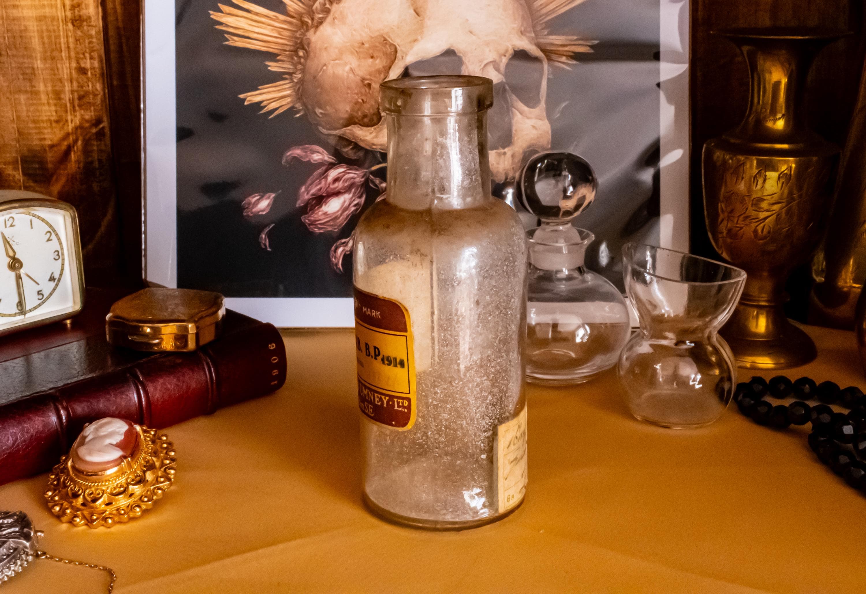 Antique clear glass apothecary bottle with a yellow and red paper label reading "Guaiacol Carb" from London, sitting next to a vintage glass stopper and an anatomical skull print.