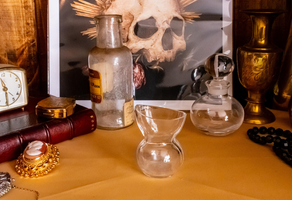 Antique clear glass apothecary bottle with a yellow and red paper label reading "Guaiacol Carb" from London, sitting next to a vintage glass stopper and an anatomical skull print.