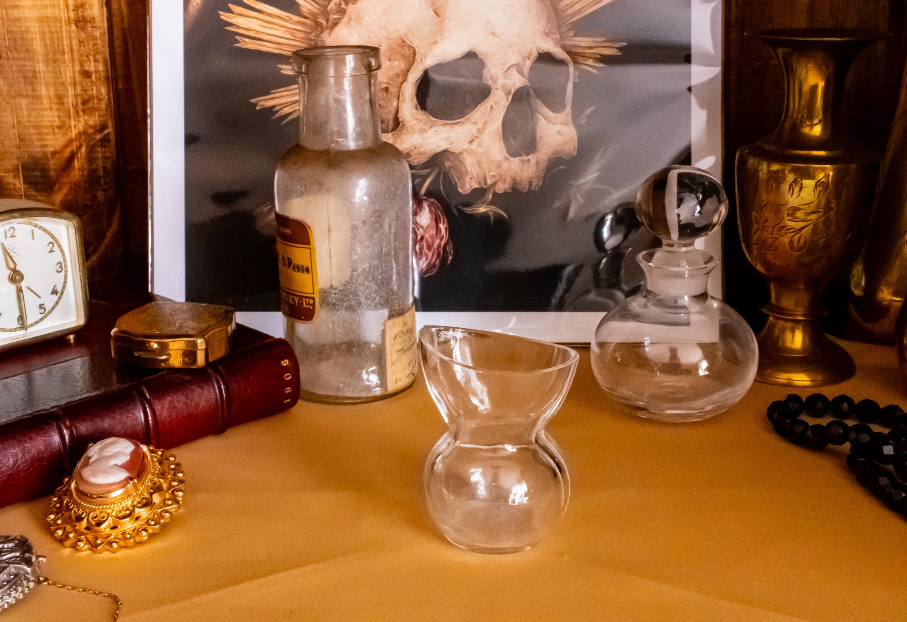 Antique clear glass apothecary bottle with a yellow and red paper label reading "Guaiacol Carb" from London, sitting next to a vintage glass stopper and an anatomical skull print.
