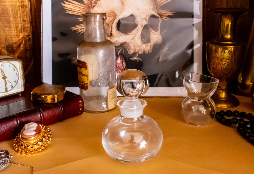 Antique clear glass apothecary bottle with a yellow and red paper label reading "Guaiacol Carb" from London, sitting next to a vintage glass stopper and an anatomical skull print.