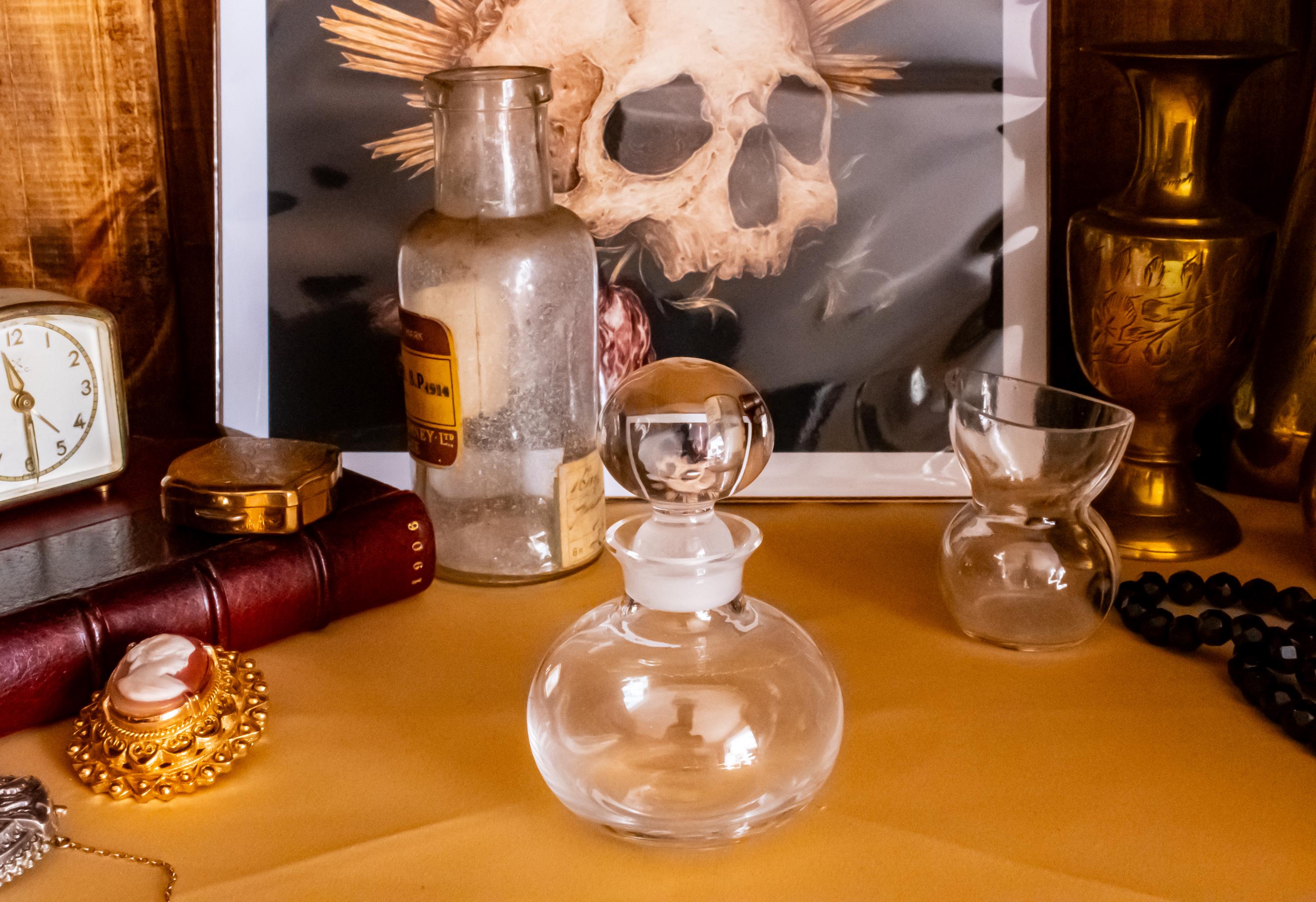 Antique clear glass apothecary bottle with a yellow and red paper label reading "Guaiacol Carb" from London, sitting next to a vintage glass stopper and an anatomical skull print.