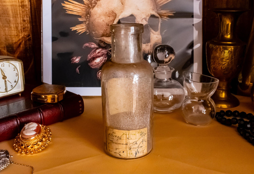 Antique clear glass apothecary bottle with a yellow and red paper label reading "Guaiacol Carb" from London, sitting next to a vintage glass stopper and an anatomical skull print.