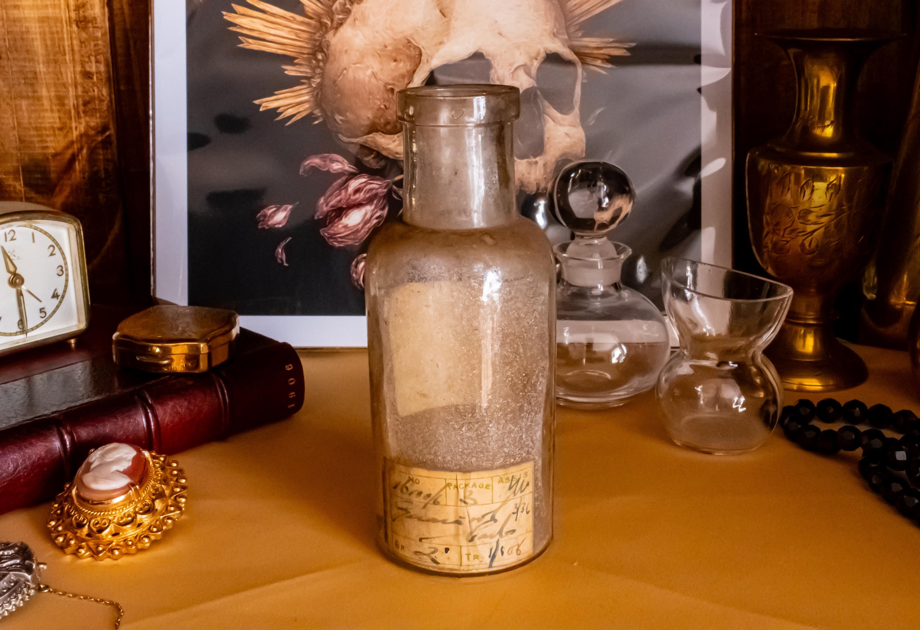 Antique clear glass apothecary bottle with a yellow and red paper label reading "Guaiacol Carb" from London, sitting next to a vintage glass stopper and an anatomical skull print.
