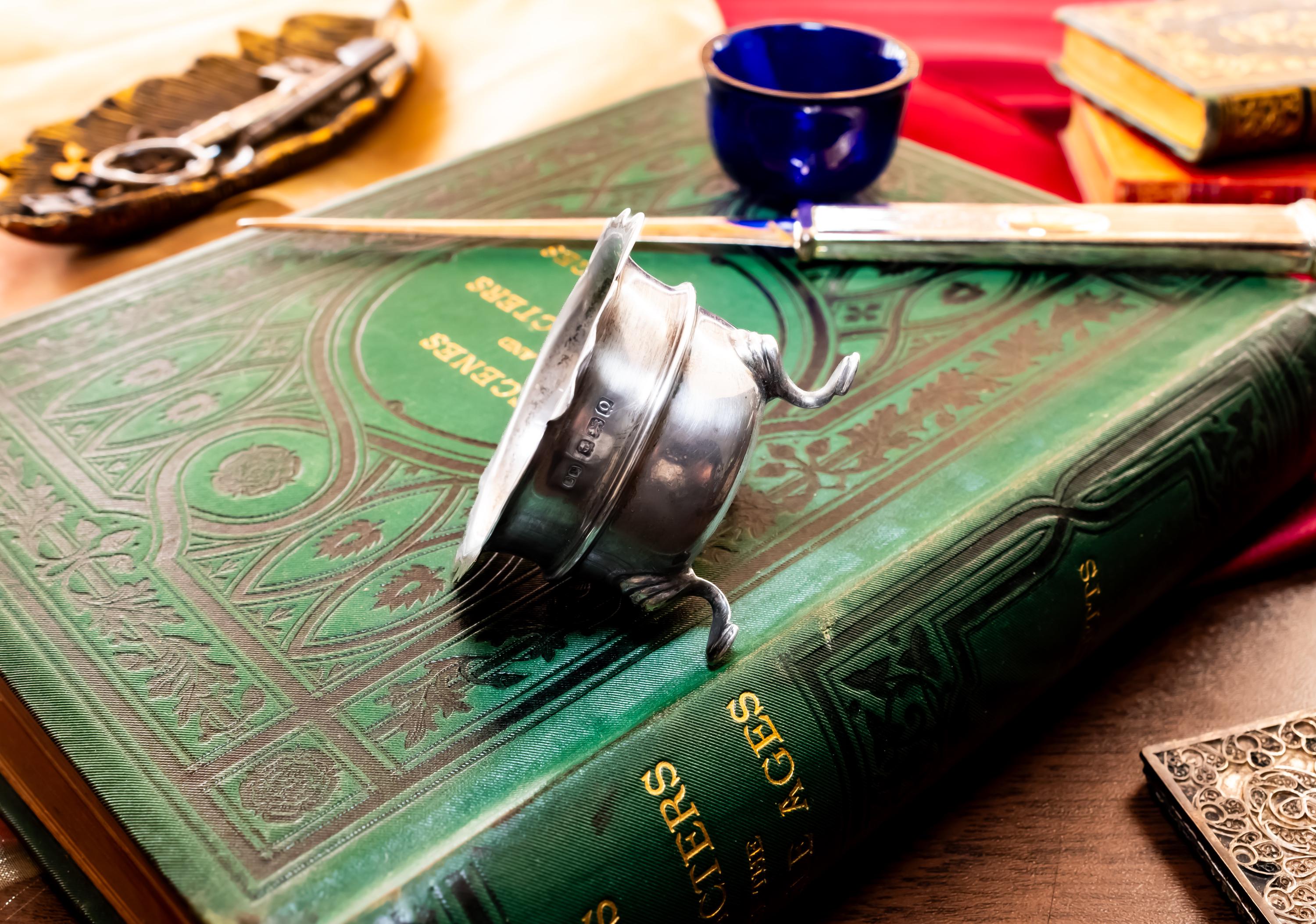Image showing the side and the stamps of Antique footed silver master salt cellar containing a vibrant cobalt blue glass liner, resting on top of an ornate green vintage book.