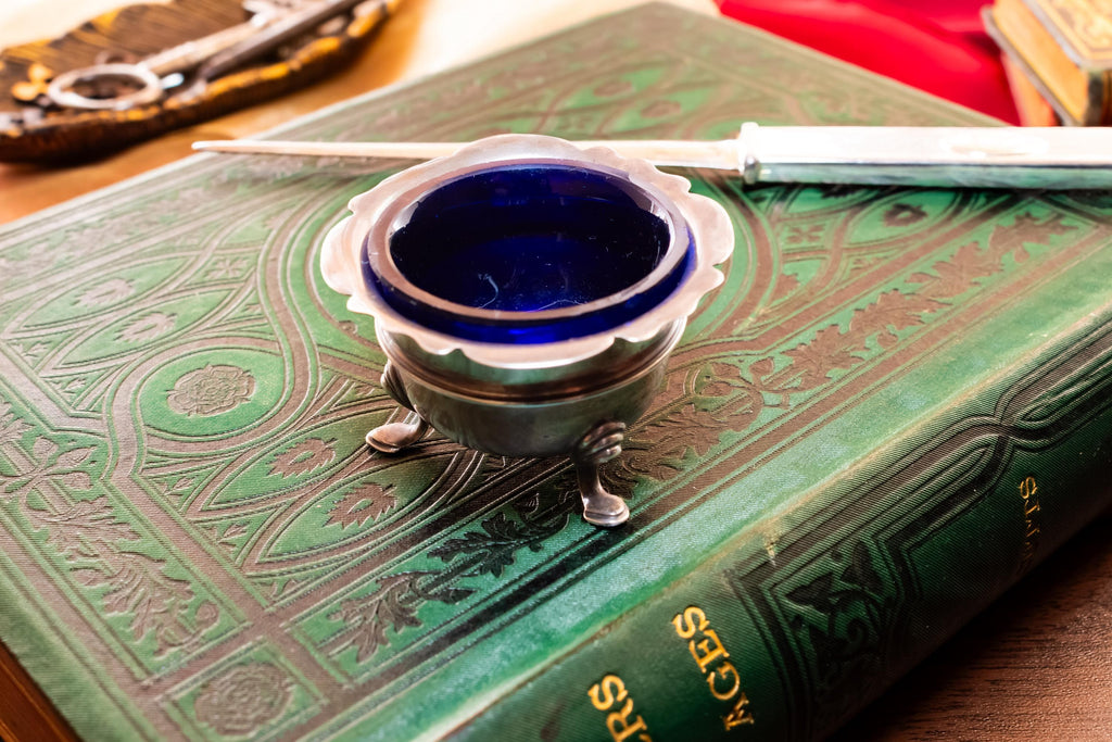 Antique footed silver master salt cellar containing a vibrant cobalt blue glass liner, resting on top of an ornate green vintage book.