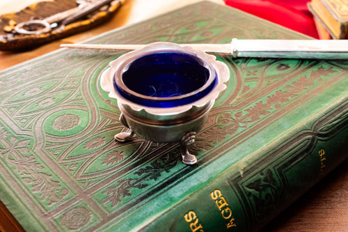 Antique footed silver master salt cellar containing a vibrant cobalt blue glass liner, resting on top of an ornate green vintage book.