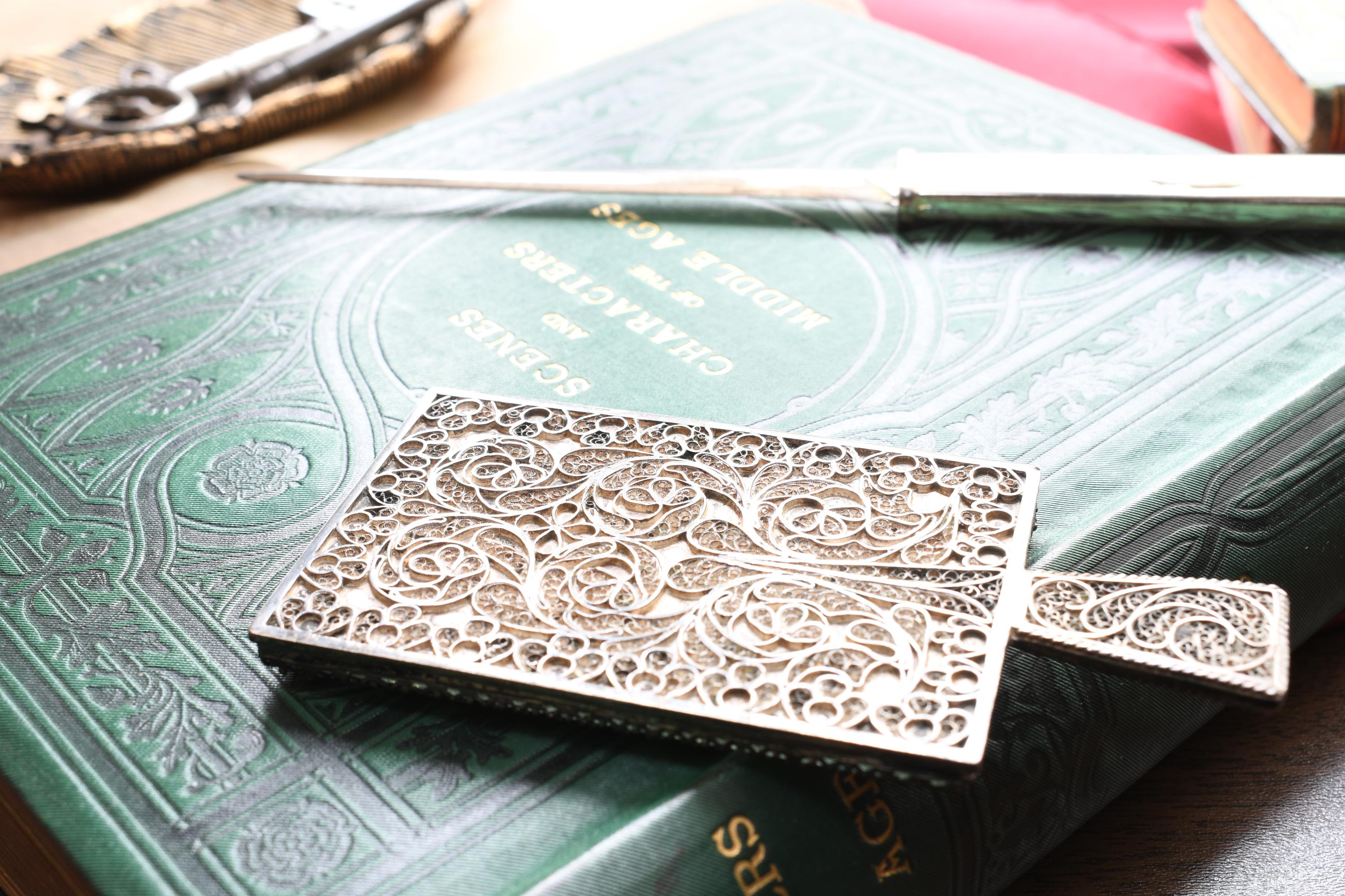 Antique rectangular silver hand mirror featuring intricate swirling filigree wirework on the back and a beveled glass front, resting on a green vintage book.