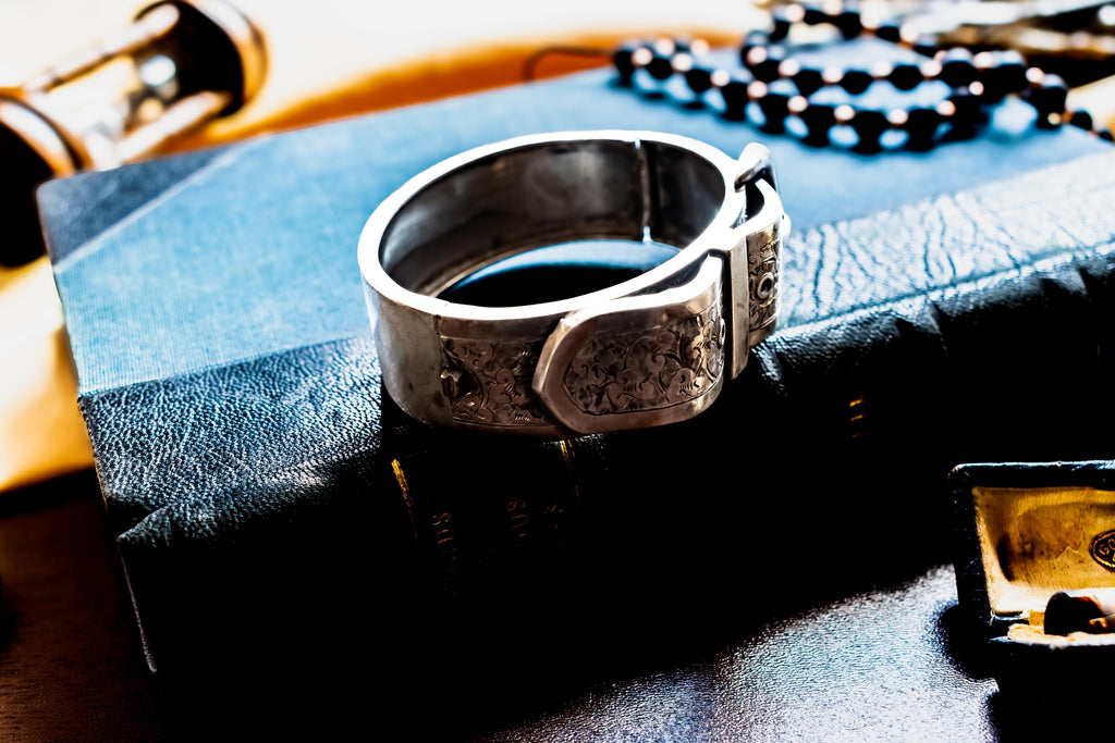 Chunky antique silver Victorian hinged bangle bracelet shaped like a buckled belt with floral engravings, resting on a dark vintage book titled Diamonds and Precious Stones.