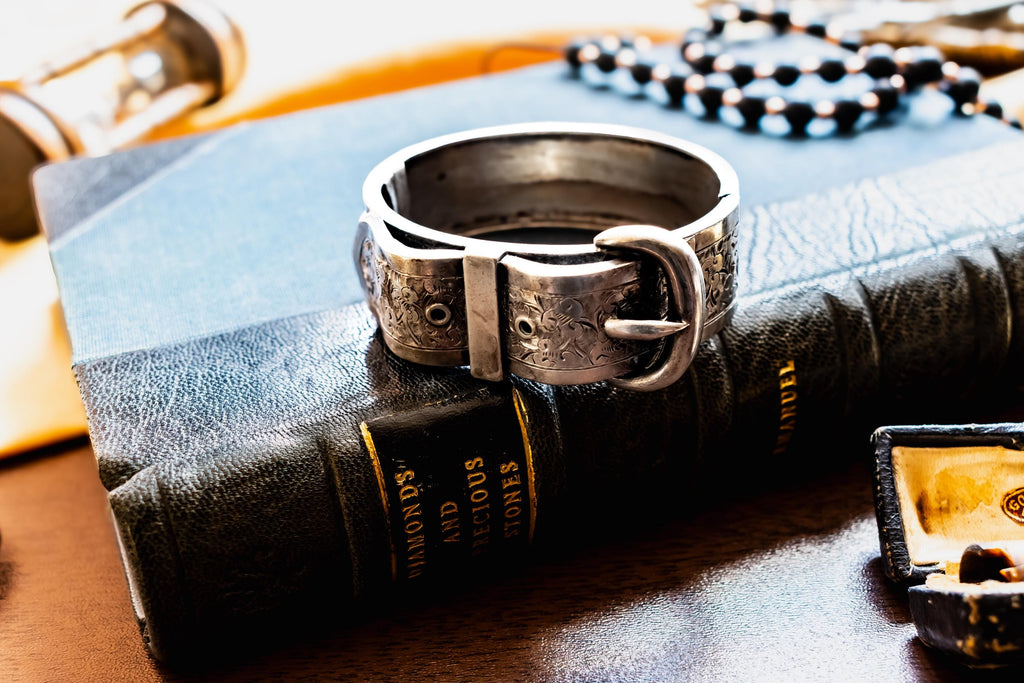 Chunky antique silver Victorian hinged bangle bracelet shaped like a buckled belt with floral engravings, resting on a dark vintage book titled Diamonds and Precious Stones.