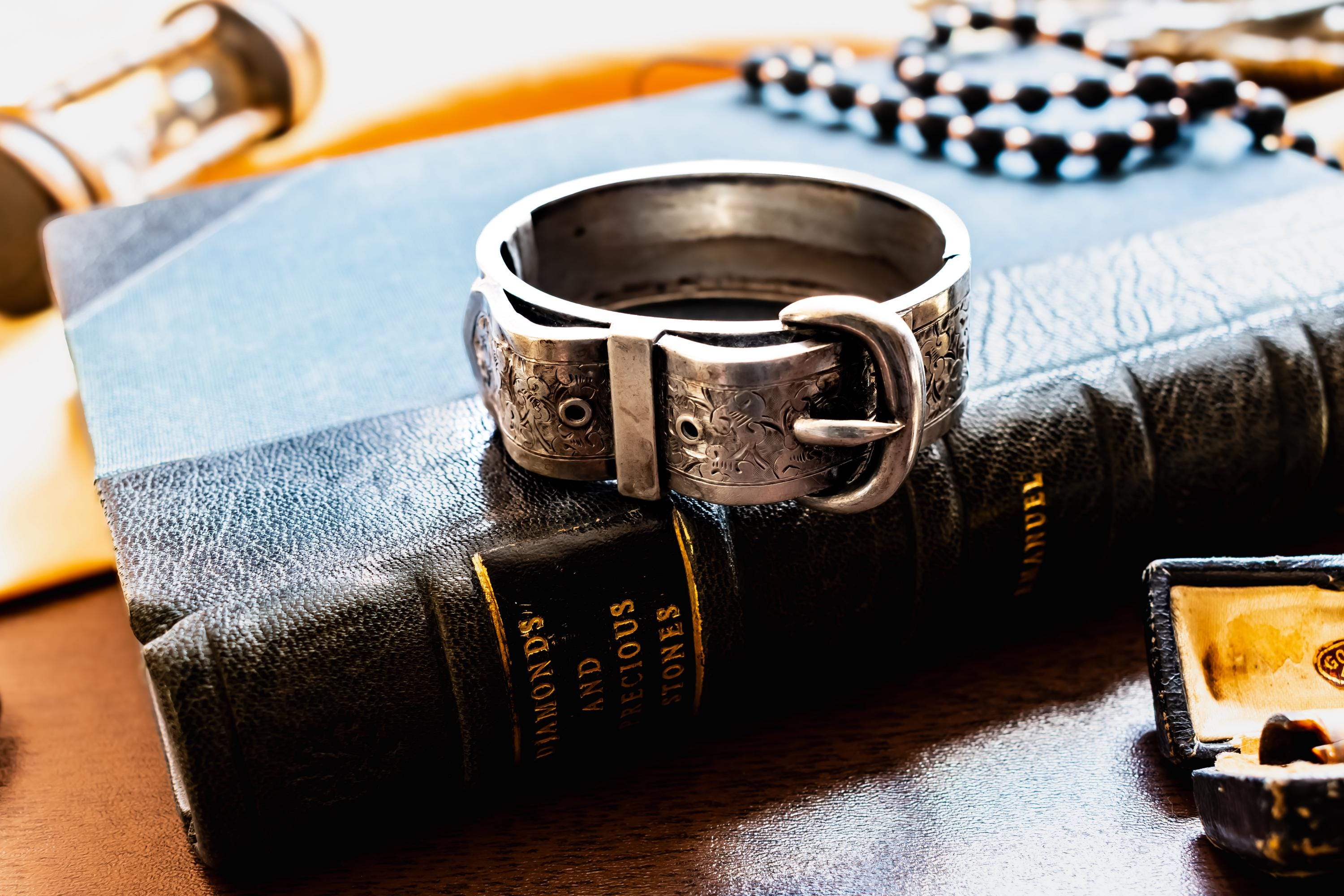 Chunky antique silver Victorian hinged bangle bracelet shaped like a buckled belt with floral engravings, resting on a dark vintage book titled Diamonds and Precious Stones.