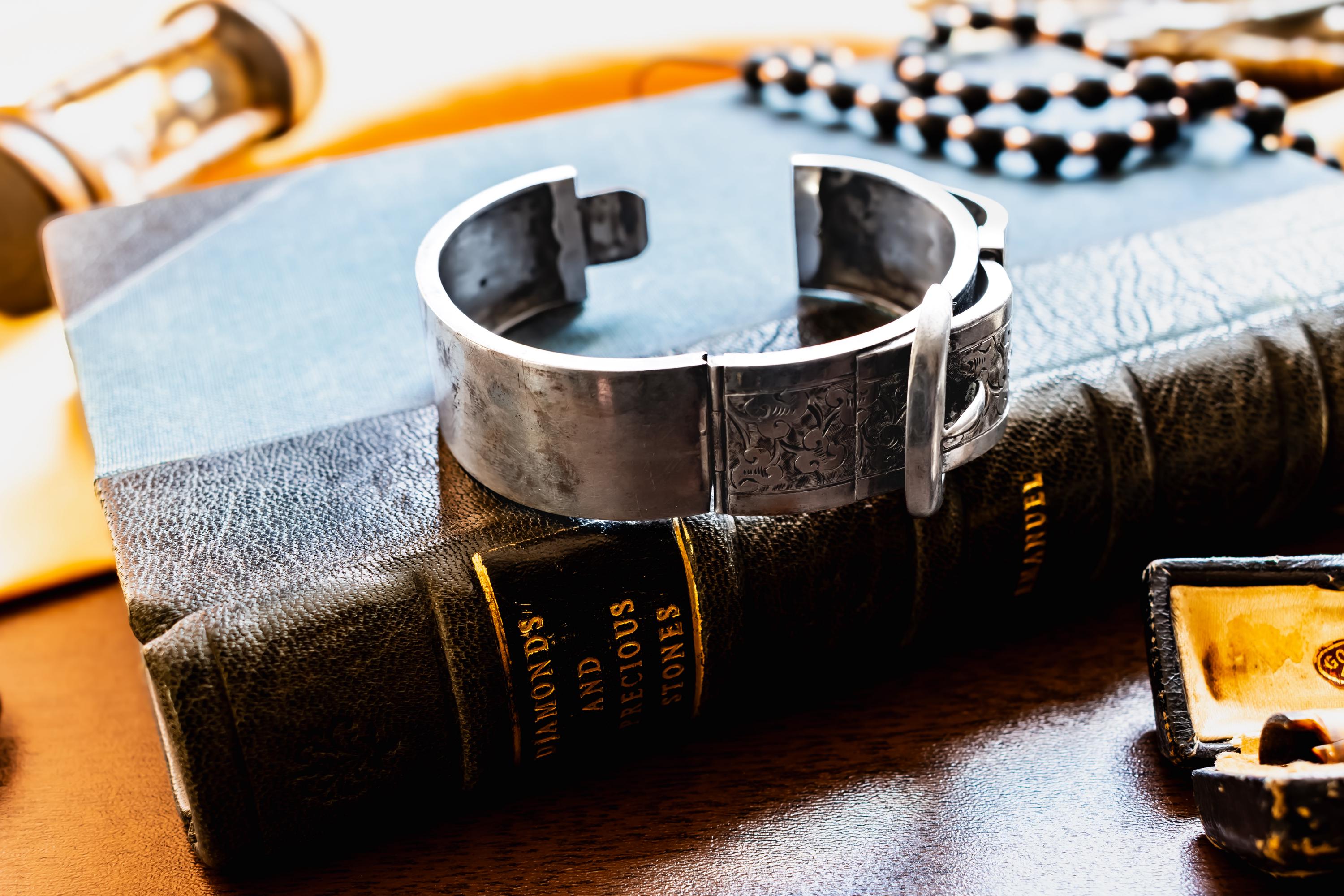 Chunky antique silver Victorian hinged bangle bracelet shaped like a buckled belt with floral engravings, resting on a dark vintage book titled Diamonds and Precious Stones.