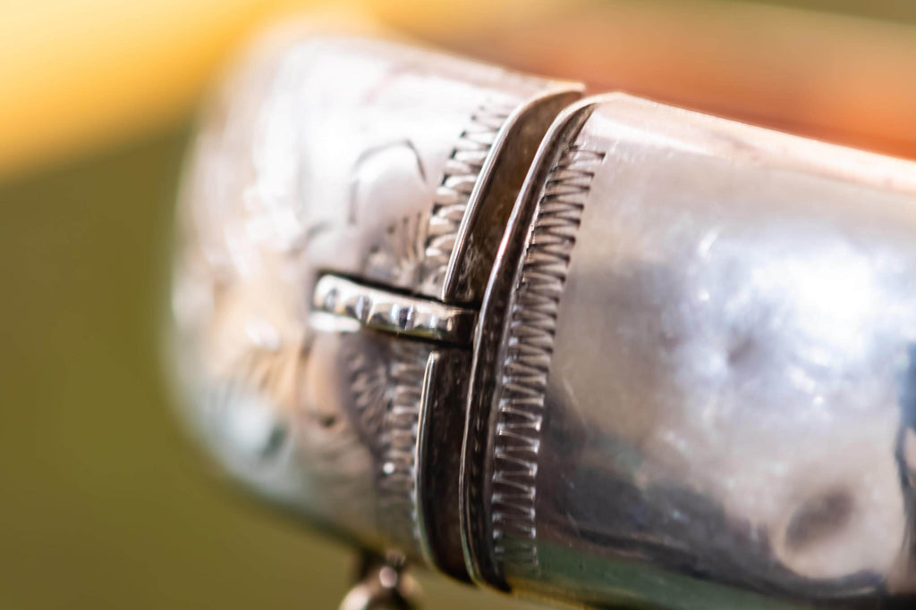Close-up of a wide, vintage 1969 Birmingham sterling silver hinged bangle with intricate botanical engravings and a safety chain, resting beautifully on a vintage leather-bound book.