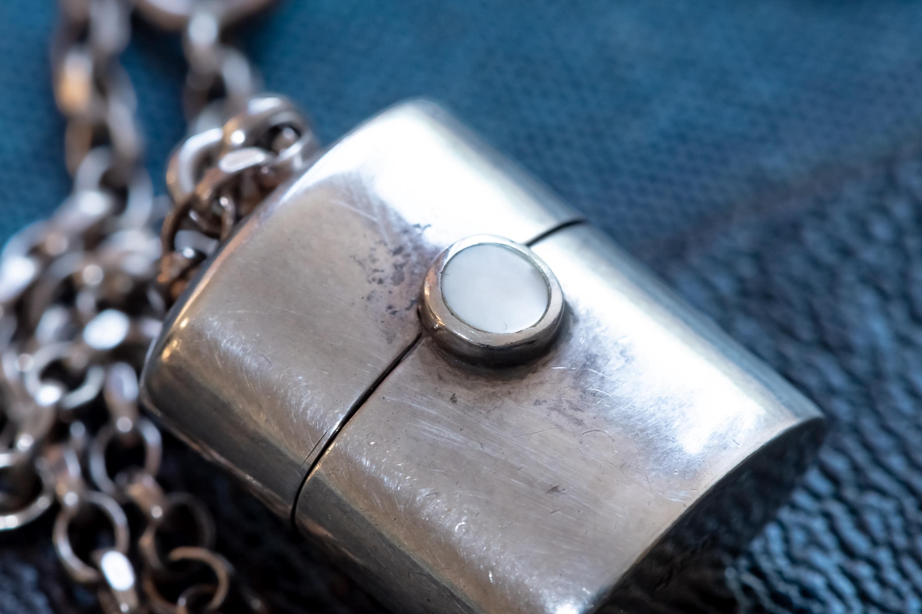 A silver-toned antique silver pill box pendant shaped like a small, rounded rectangular container with a hinged lid. The centre of the lid features a circular white inlay. The pendant is attached to a thick, textured silver-toned metal chain link necklace.