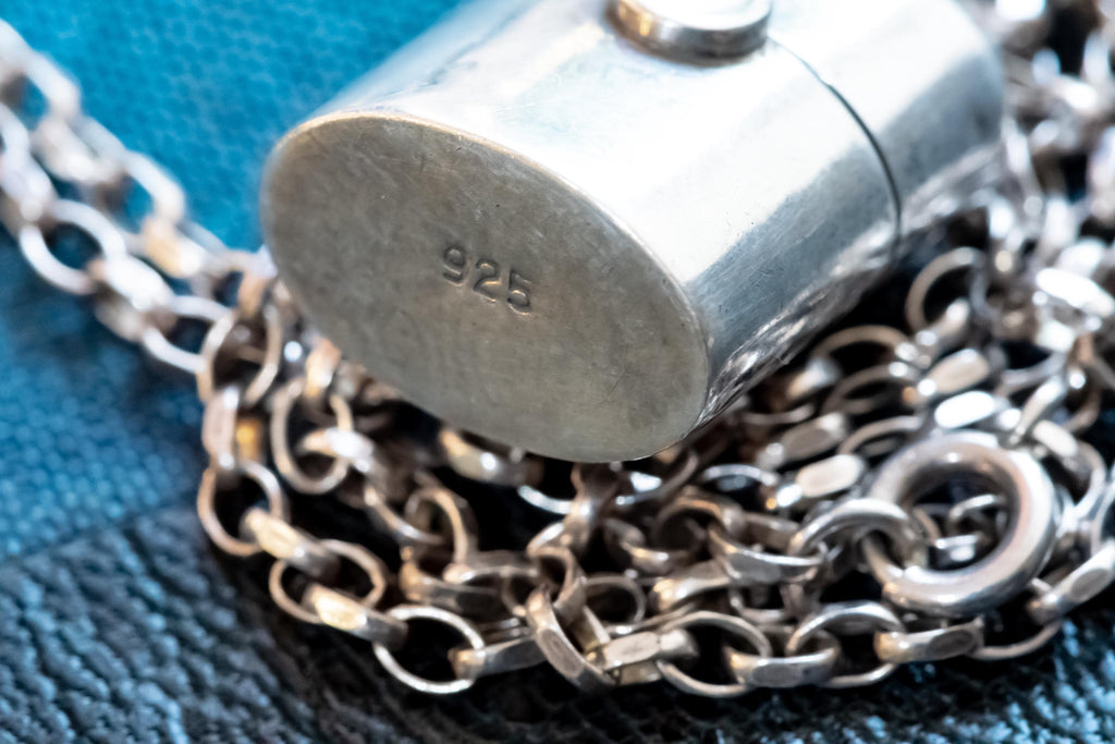A silver-coloured antique silver pill box pendant with an oval base stamped with the number 925, attached to a linked metal chain. The pendant has a smooth, reflective surface and is positioned resting on top of the coiled chain links.
