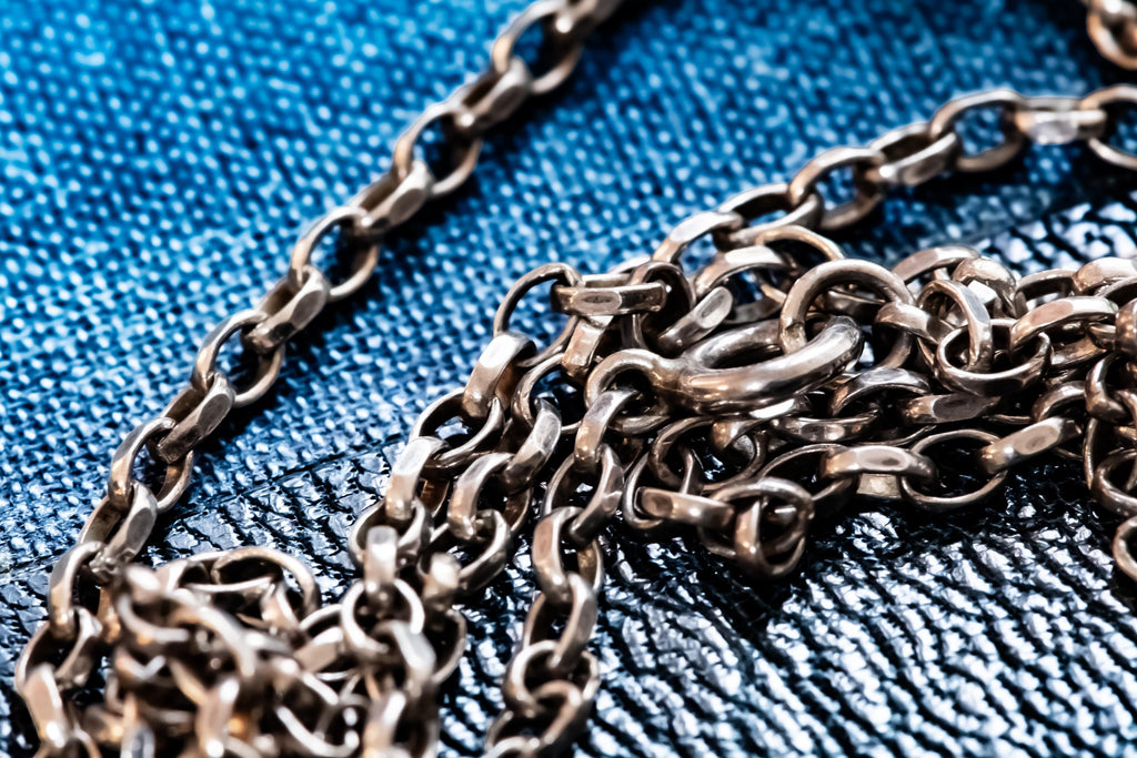 A silver metallic chain necklace featuring interconnected oval links with a polished finish. The chain is arranged in a loose, coiled formation, showcasing the uniform shape and reflective surface of each individual metal link in the design.