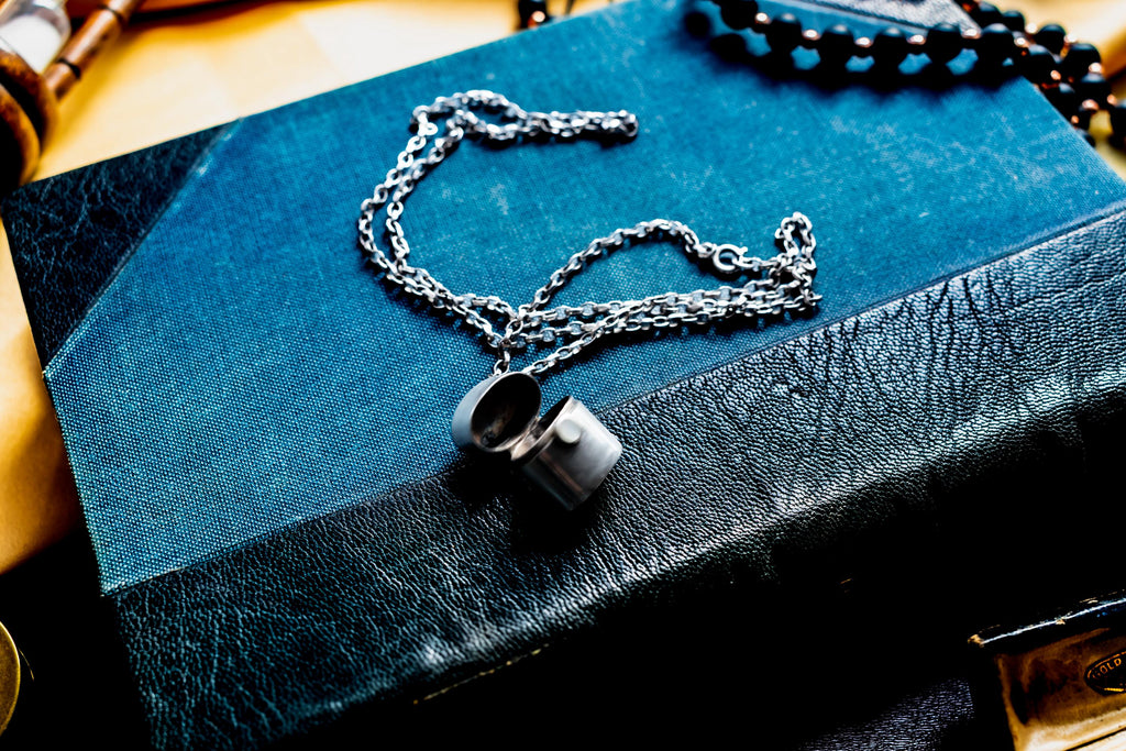 A silver-toned antique silver pill box pendant on a matching silver chain, resting on a dark blue and black textured book cover. The locket is open, revealing a hollow interior, and the chain is draped in a loose, circular pattern across the surface.